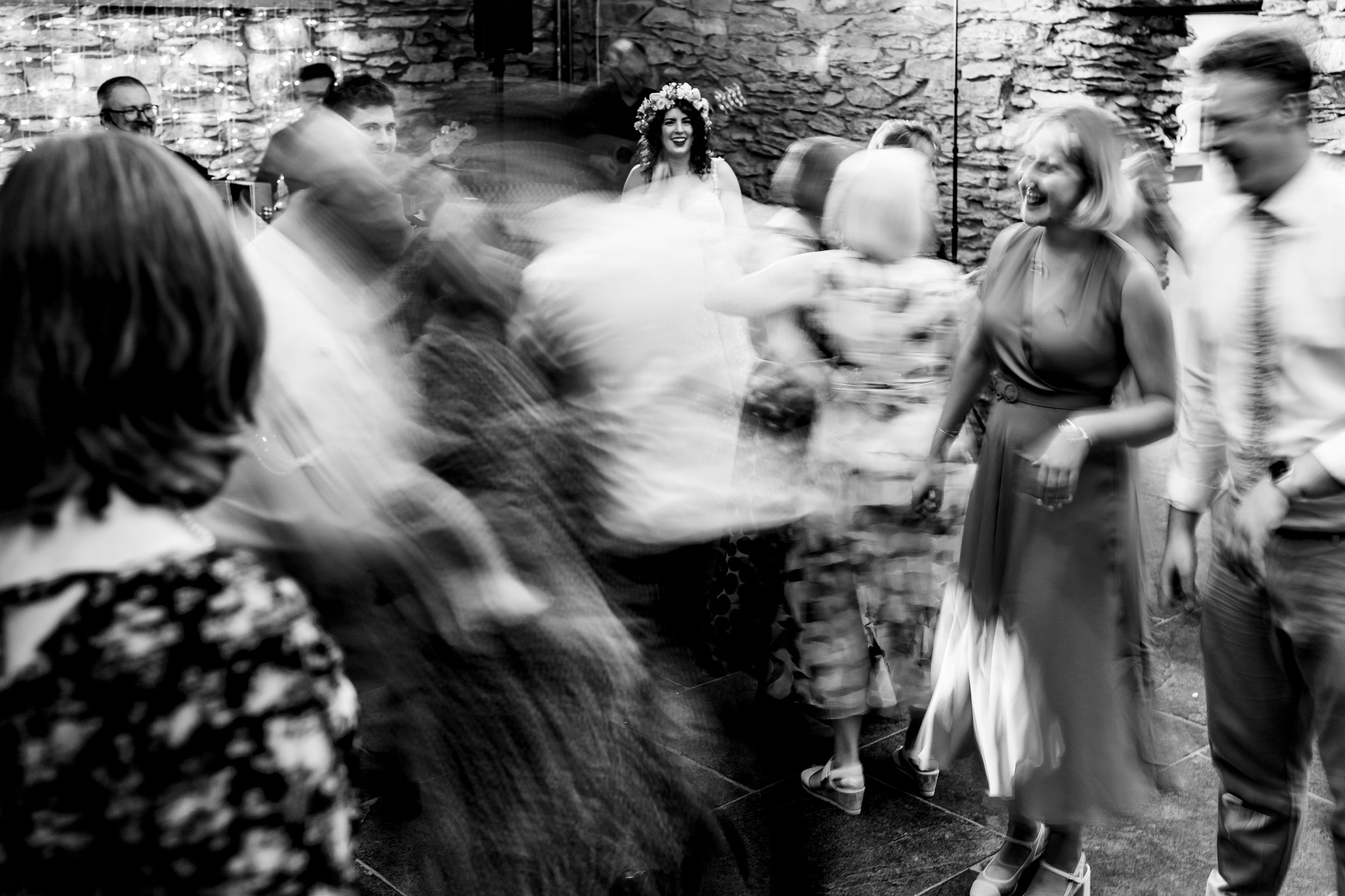 Black and white photo of bride in floral crown dancing with guests at lively indoor wedding celebration at Grasmere