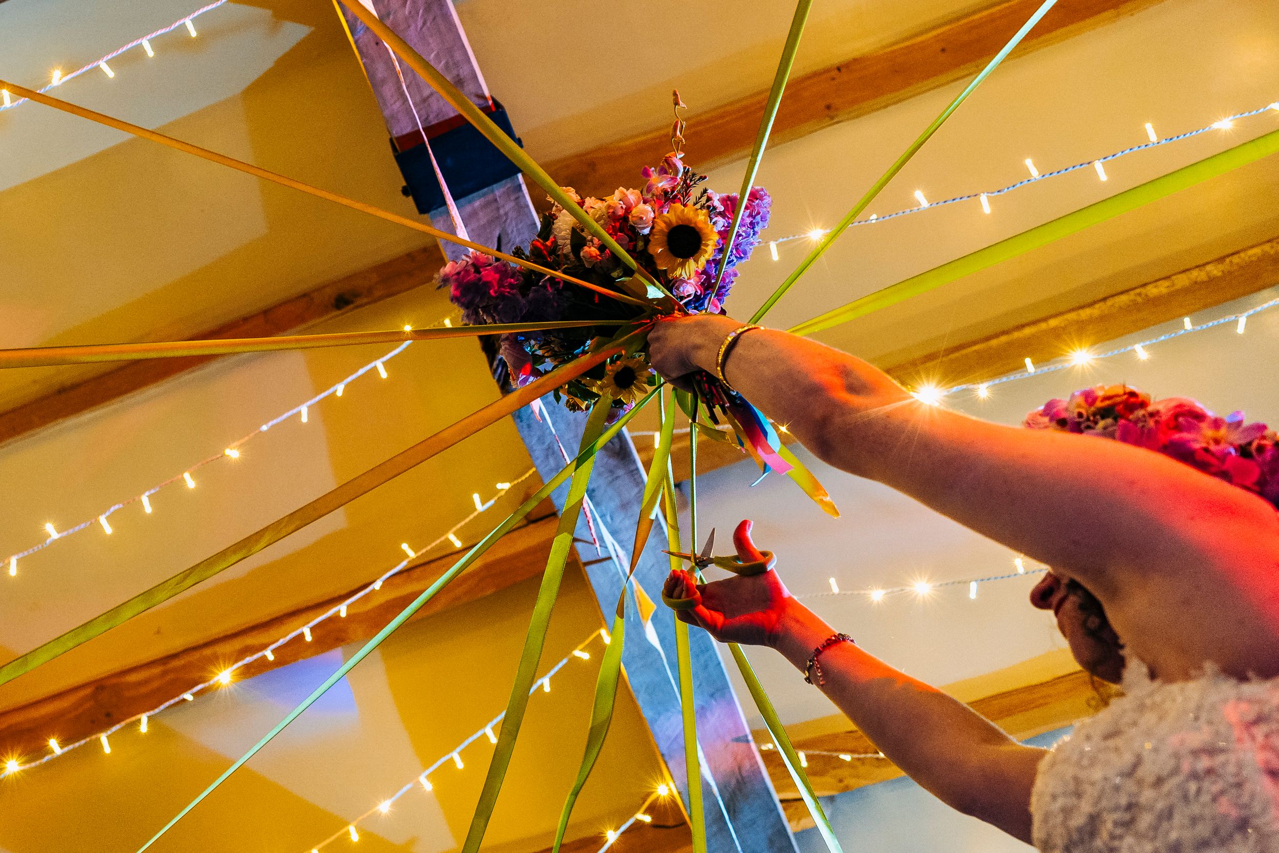 Close-up of hands holding colorful floral bouquet with ribbons under fairy lights at wedding reception.