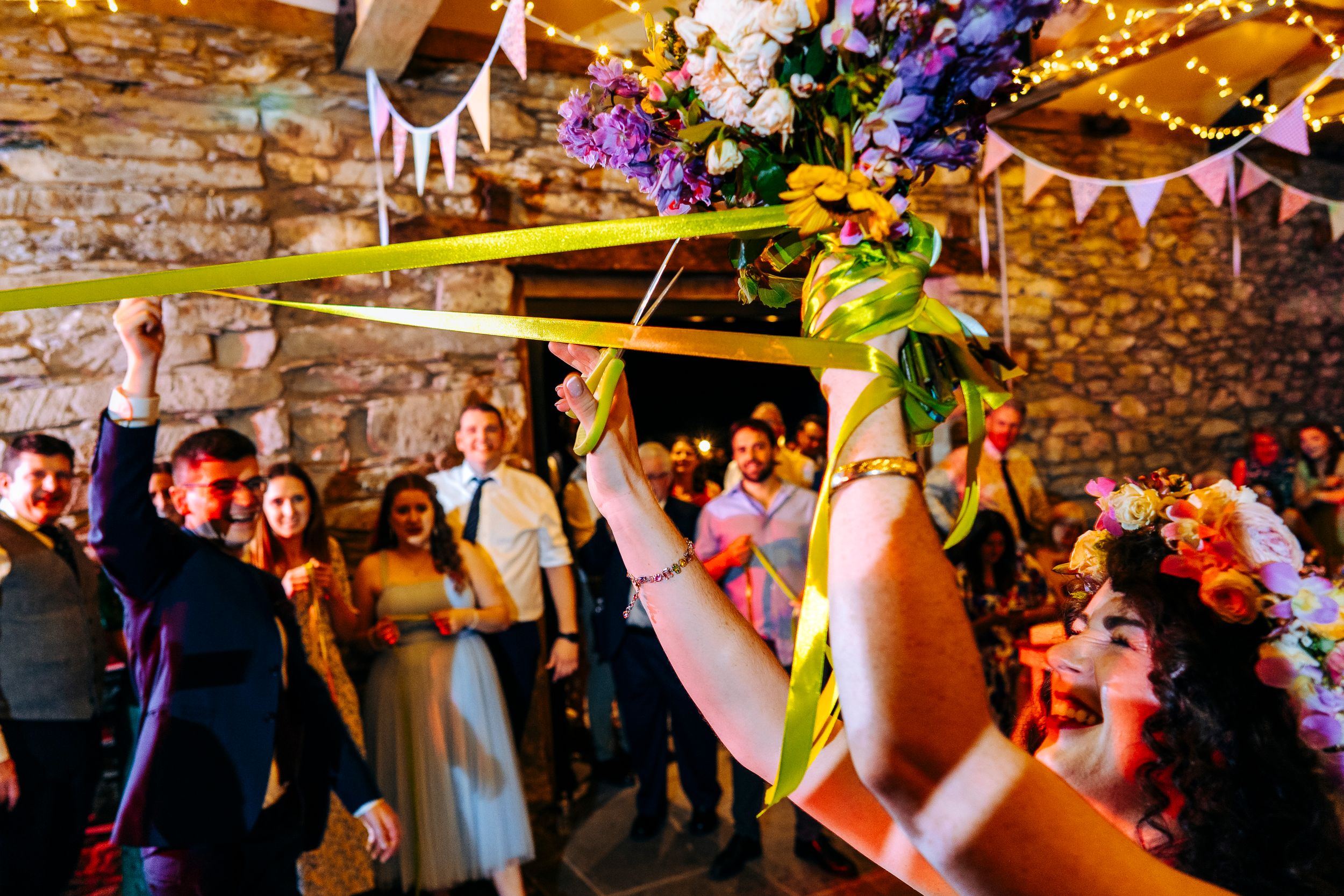 Wedding guests cheering as bride holds floral bouquet aloft in rustic stone barn decorated with bunting.