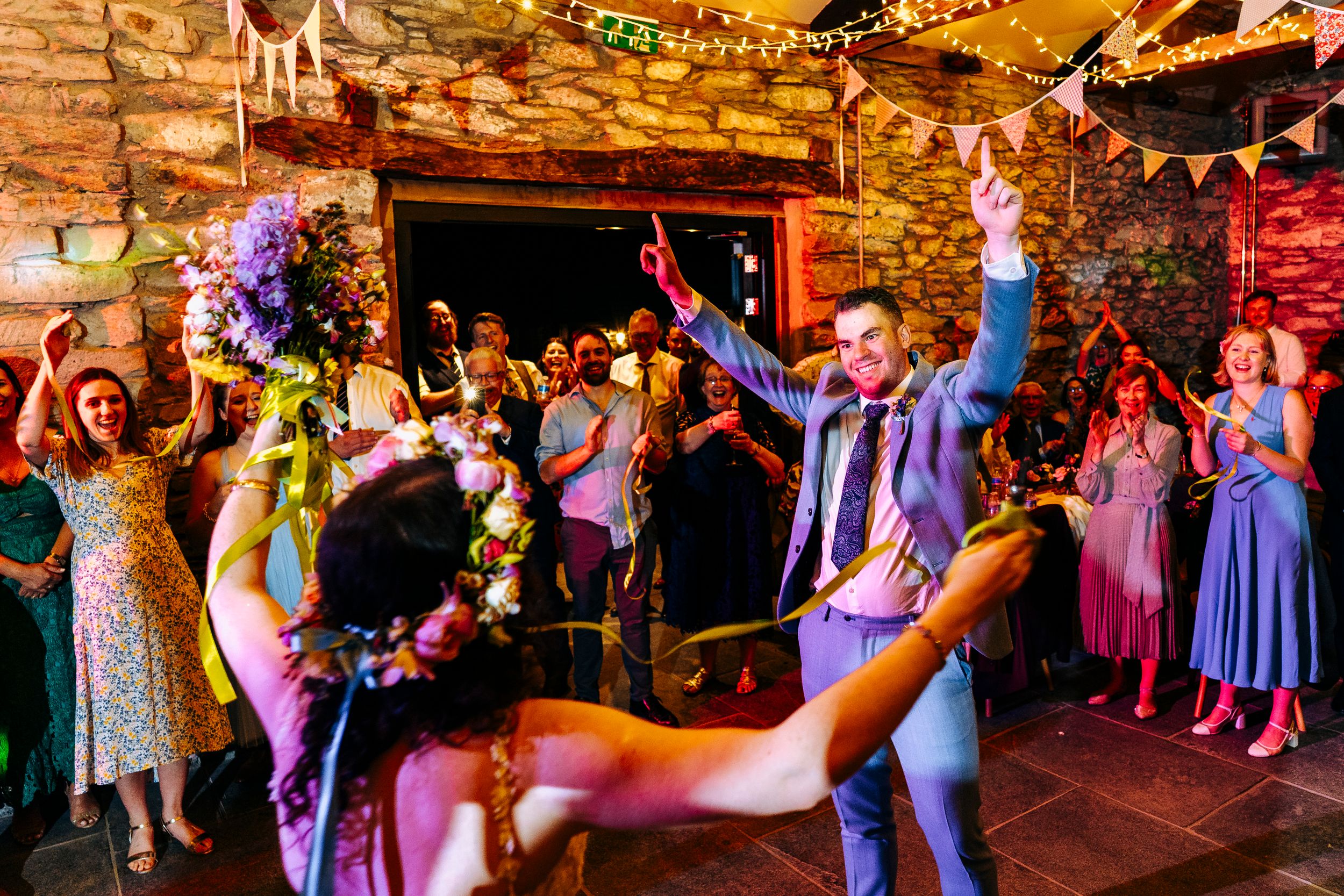 Bride and groom dancing energetically with cheering guests in decorated rustic stone barn wedding venue.