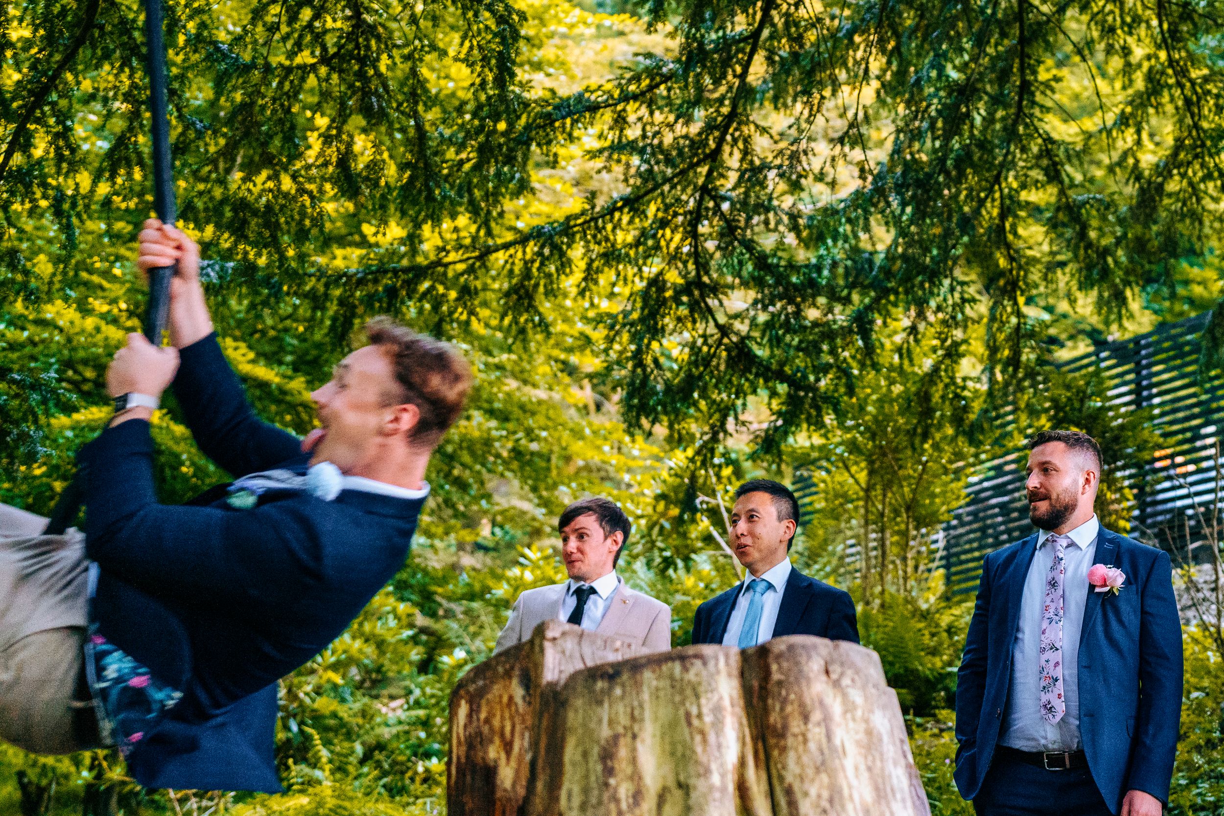 Groomsmen laugh outdoors near a tree stump as one man swings on a rope in a lush green forest setting.