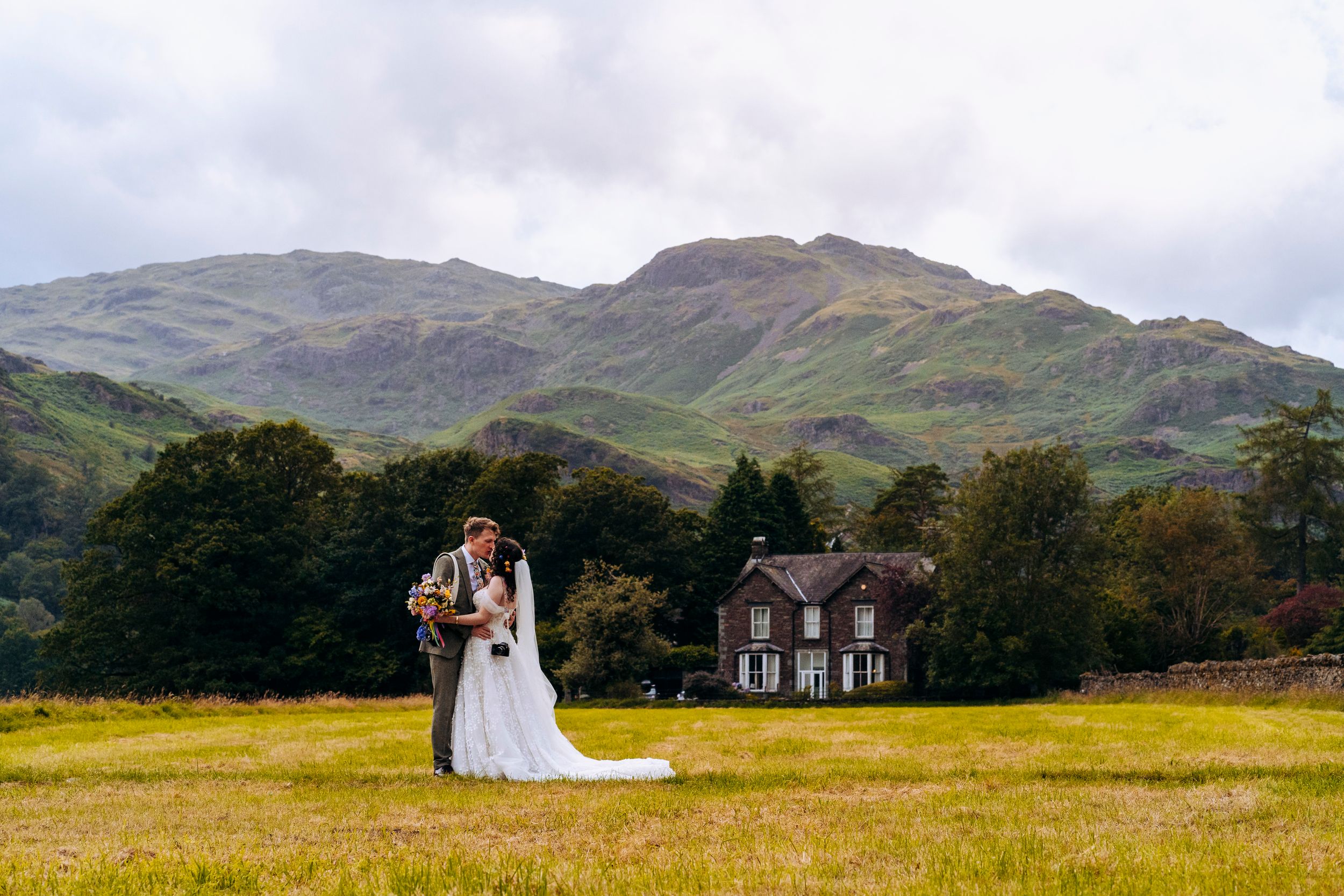 A bride and groom embrace in a vast green field with a stunning mountain backdrop and a country house behind them In Grasmere