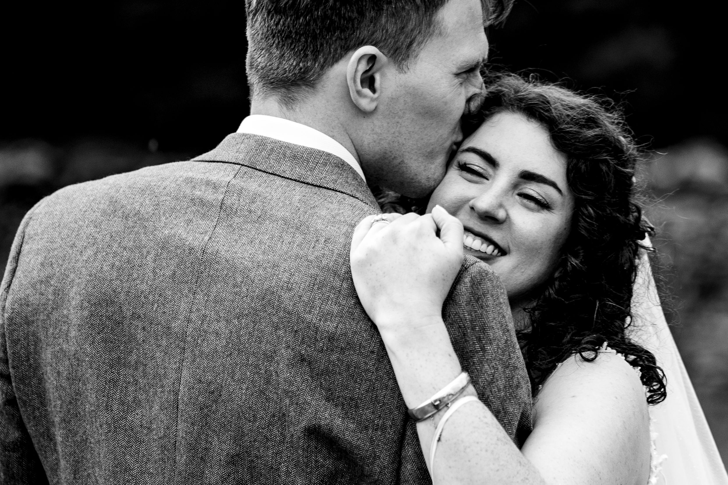 Black and white photo of a smiling bride touching her lips as the groom kisses her cheek at their wedding.
