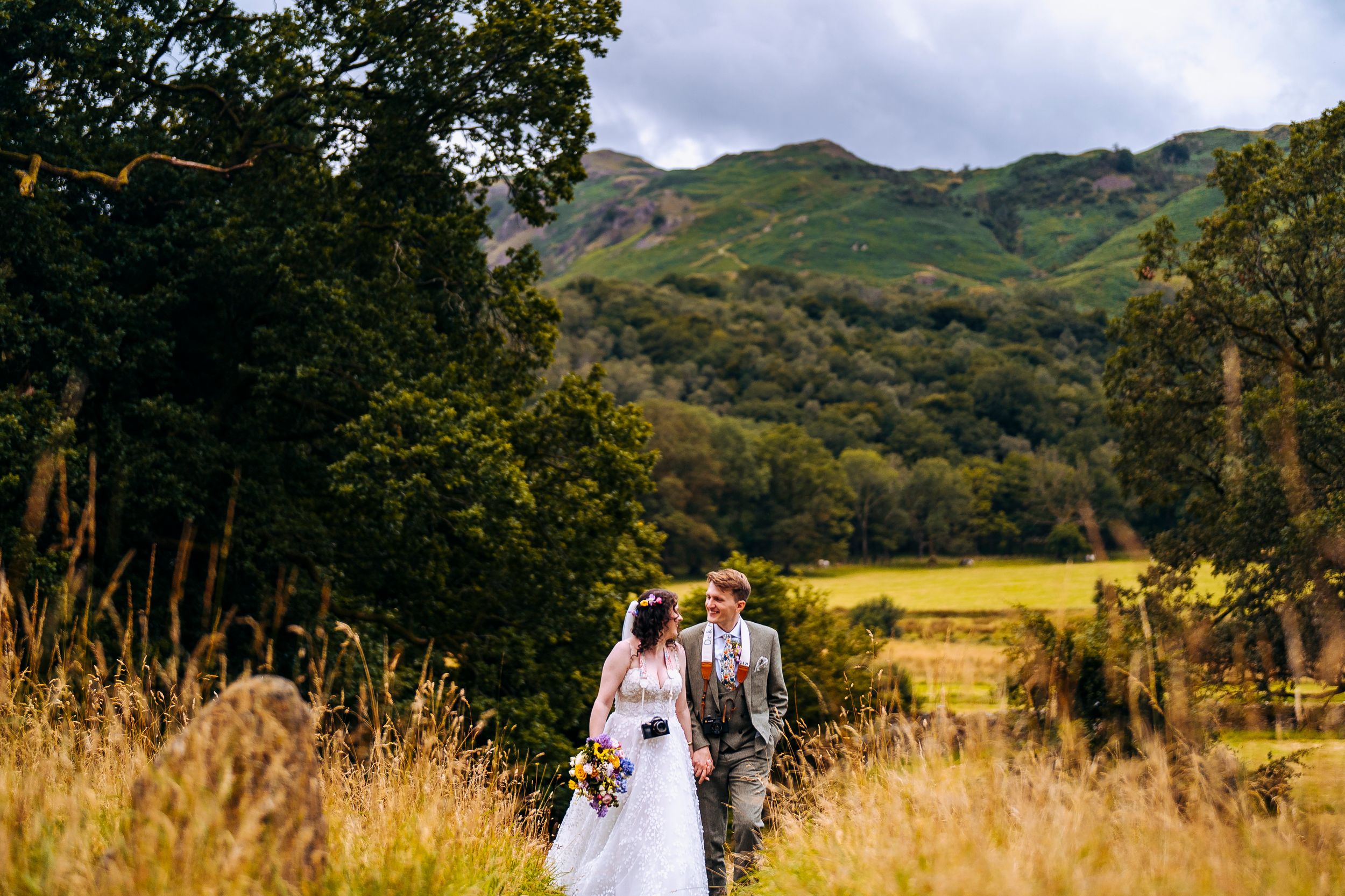Bride and groom walking at the lancrigg with mountain backdrops while they carry their cameras to photograph it too