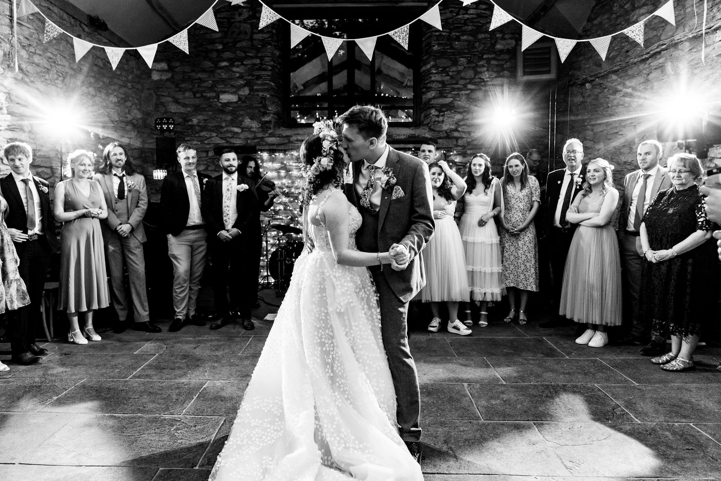 Black and white photo of a bride and groom sharing their first dance in a rustic barn with guests watching.
