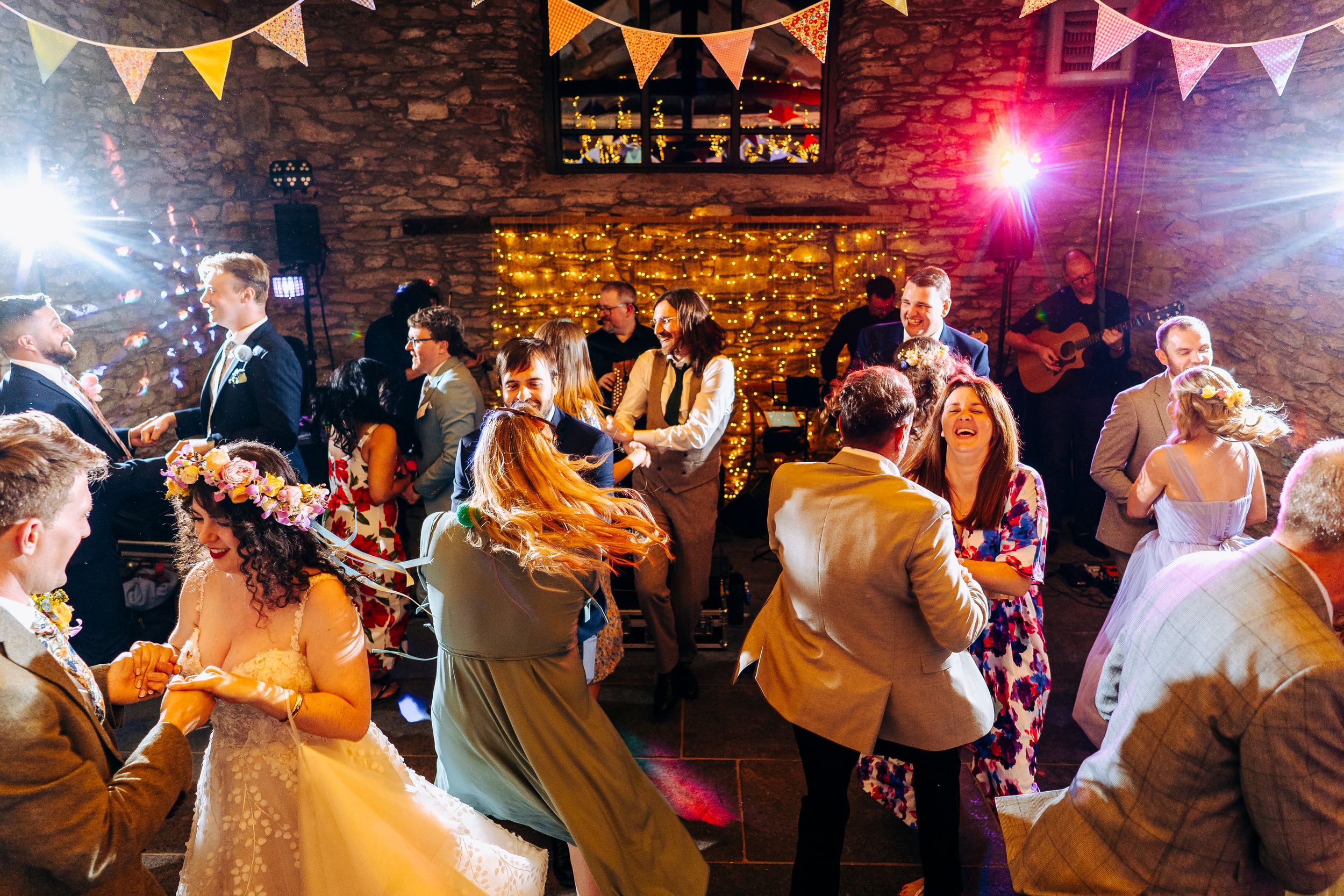 Wedding guests dance energetically in a warmly lit rustic stone barn decorated with bunting and string lights.