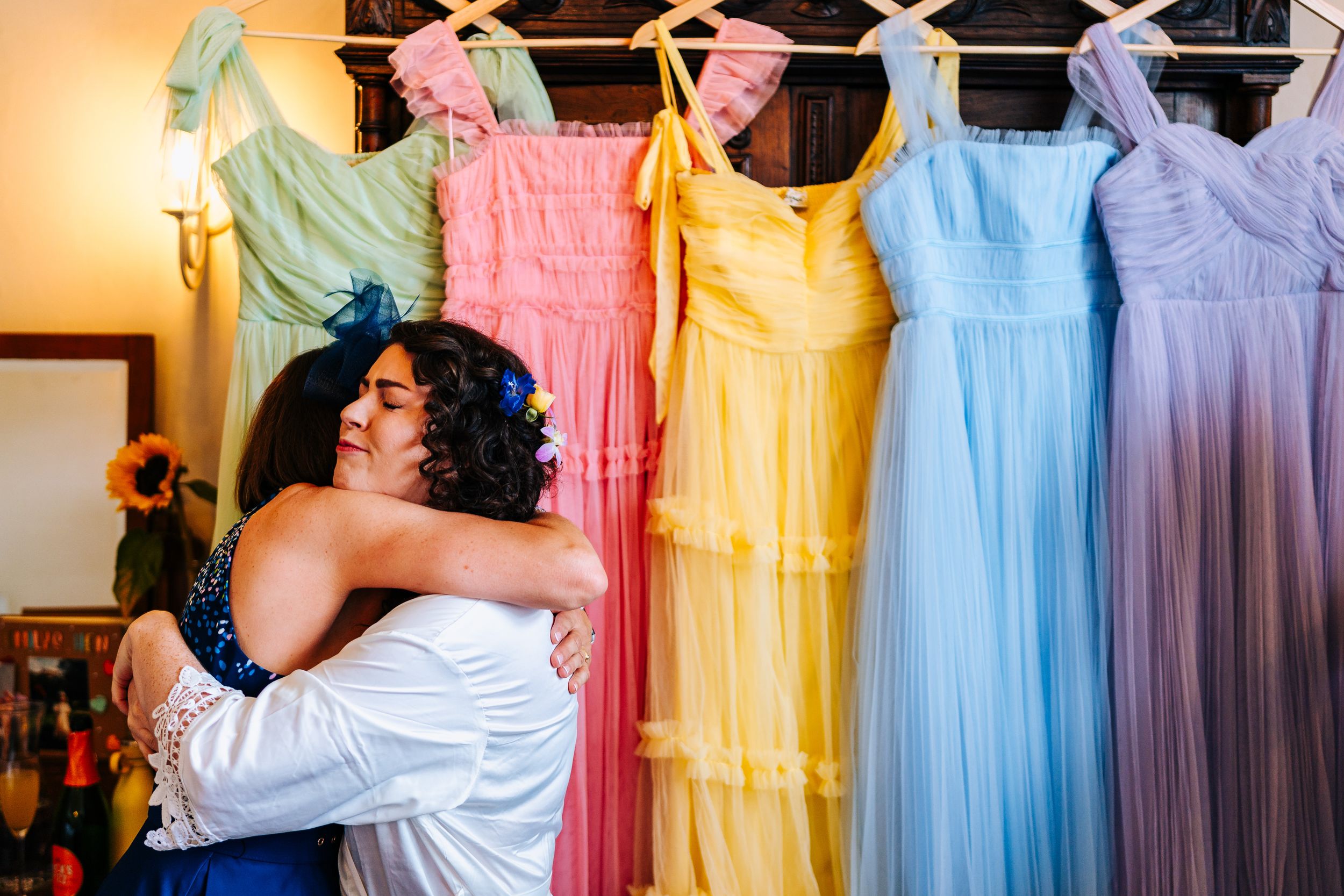 Bride embracing a guest in front of colorful pastel bridesmaid dresses on hangers.