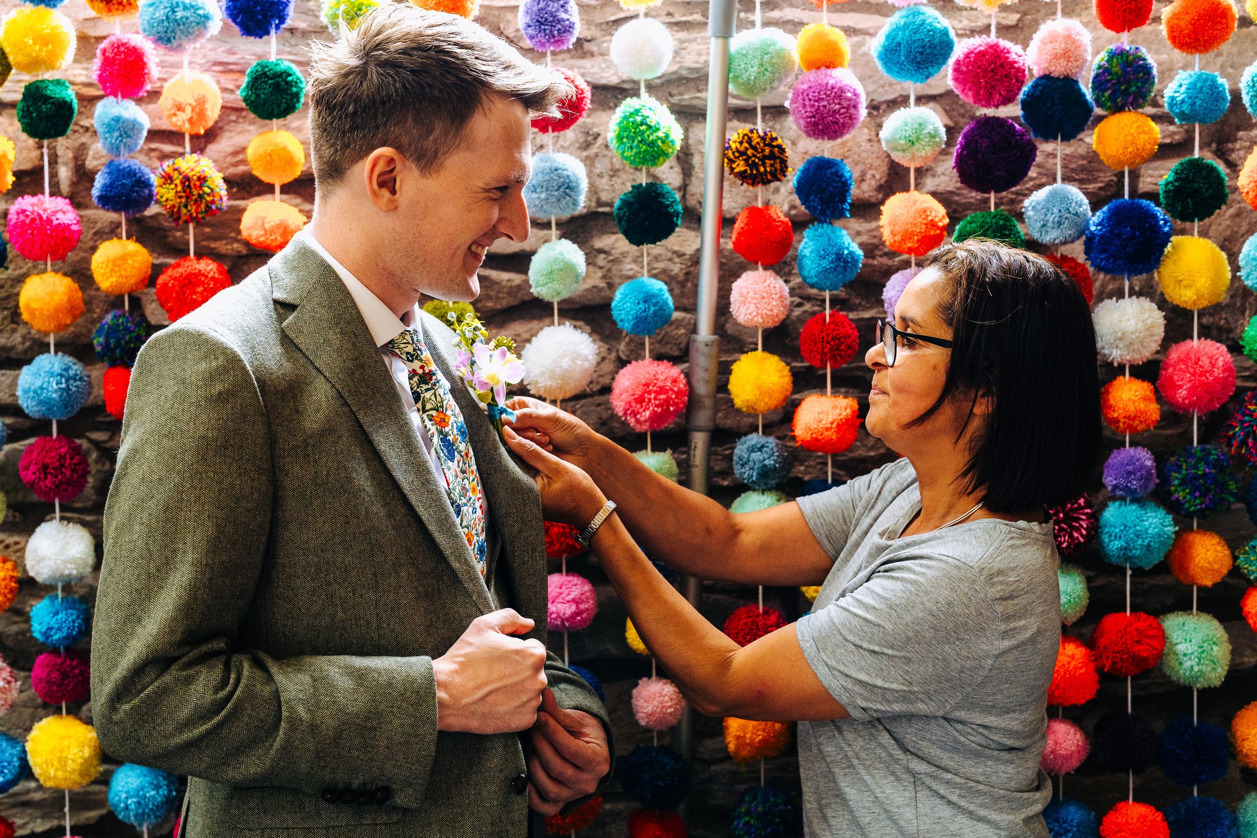 Woman pinning a floral boutonniere on a groom's lapel against a colorful pom-pom backdrop.