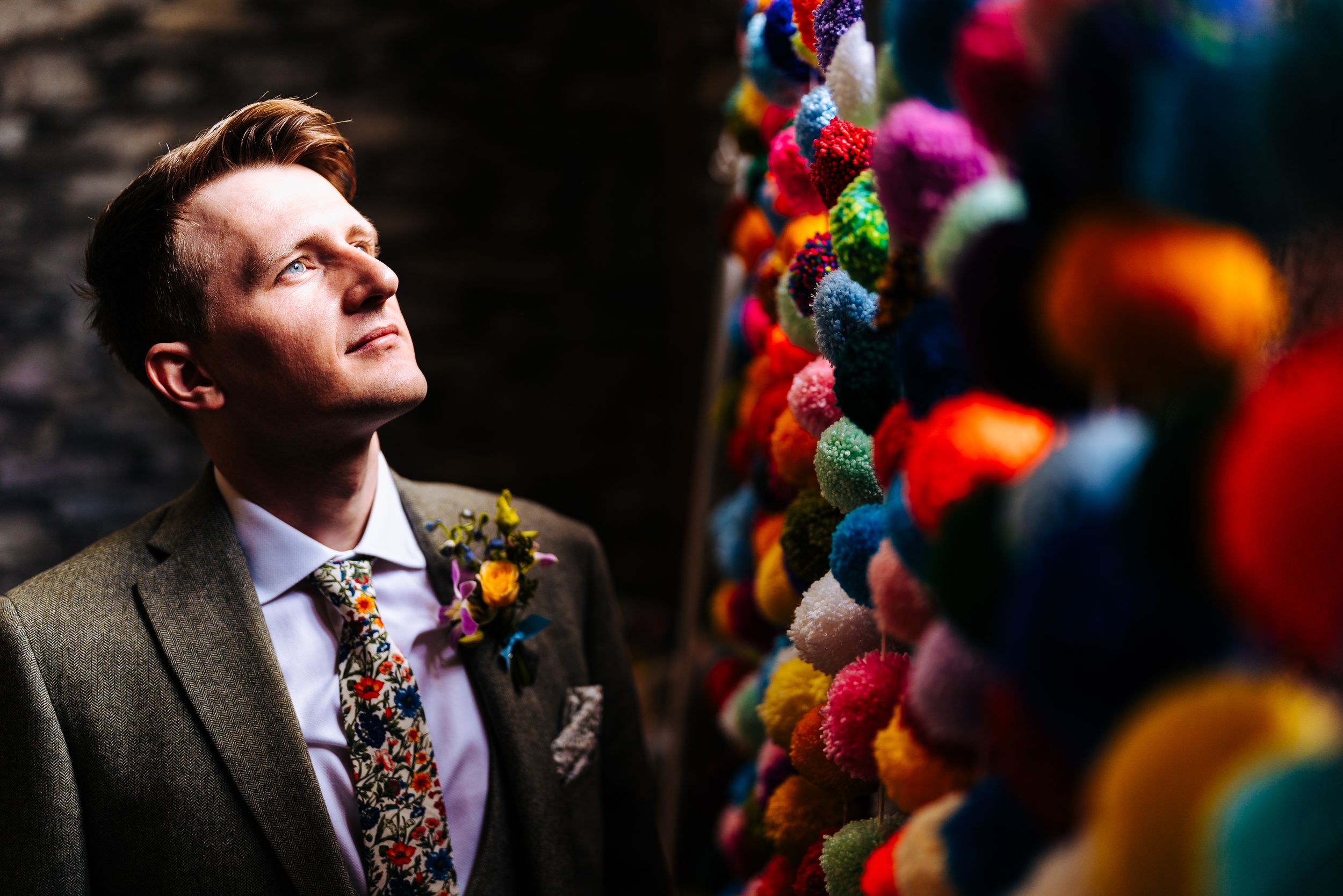 Dapper groom in a tweed suit with a floral tie standing beside colorful pom-pom decorations.