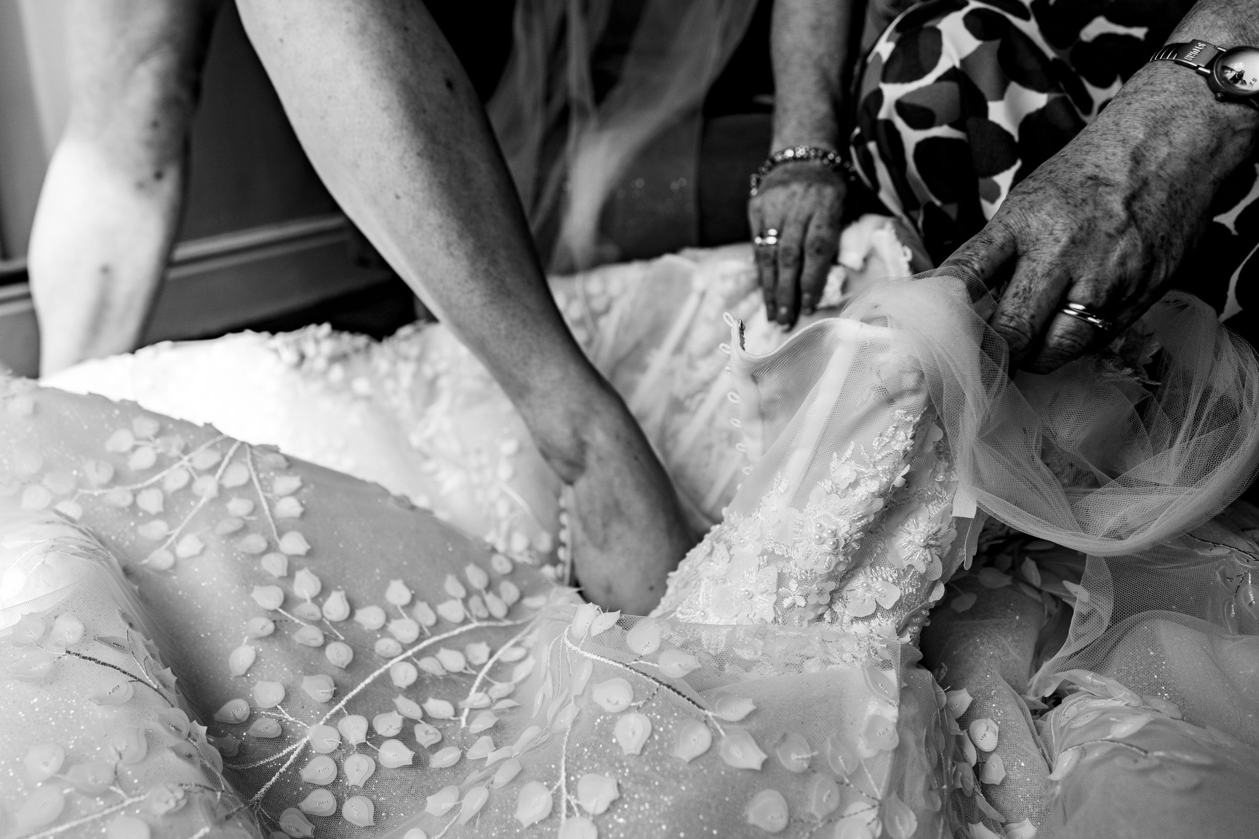 Close-up black and white photo of hands adjusting intricate floral lace wedding dress details.