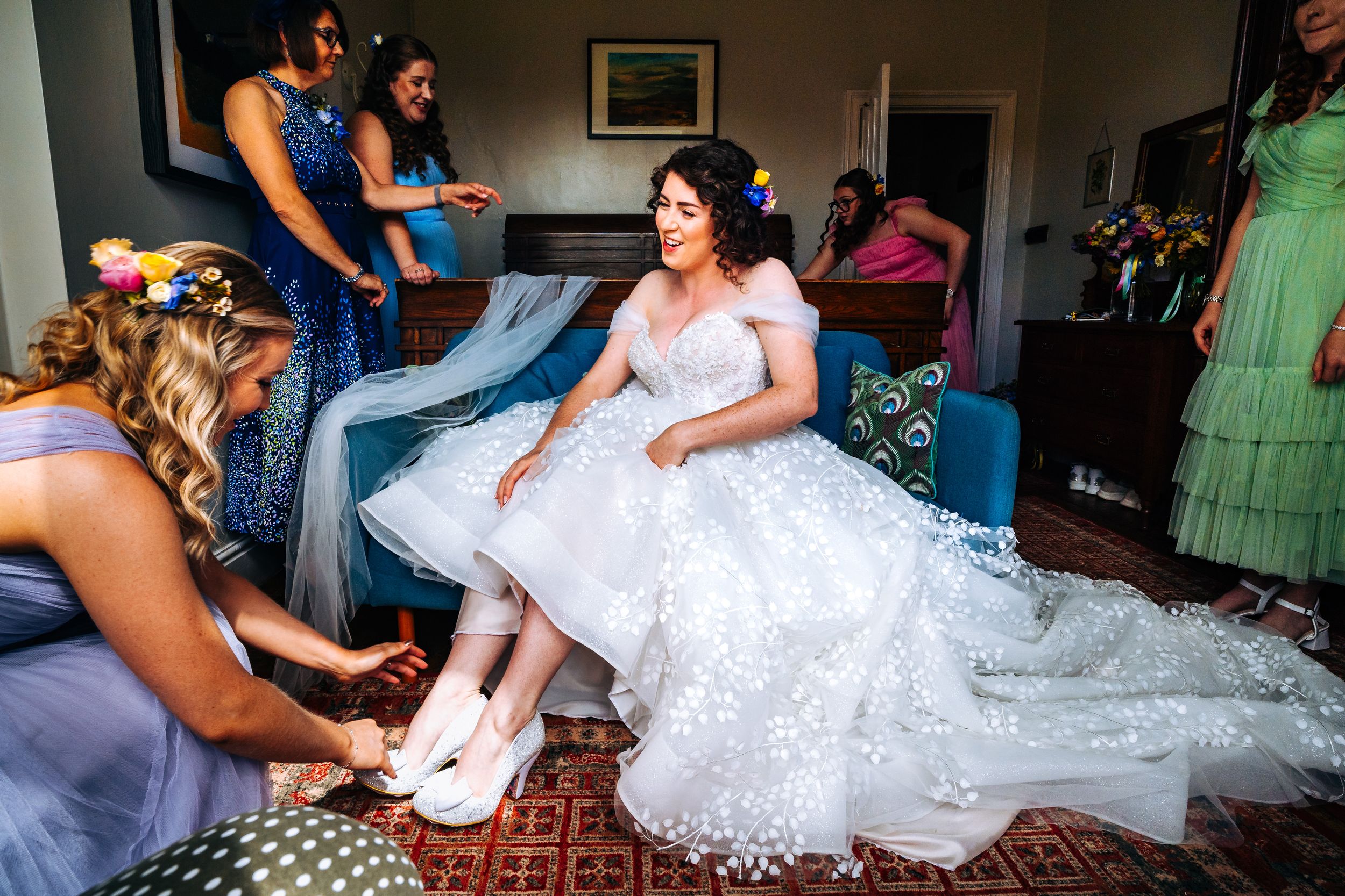 Bridesmaids in colorful dresses help bride put on wedding shoes in elegant room.
