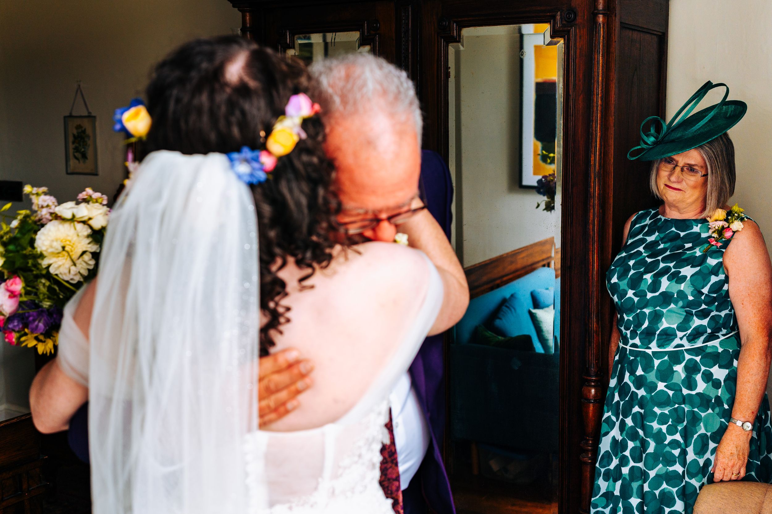 Father embraces bride in wedding dress while mother watches warmly in mirror reflection.