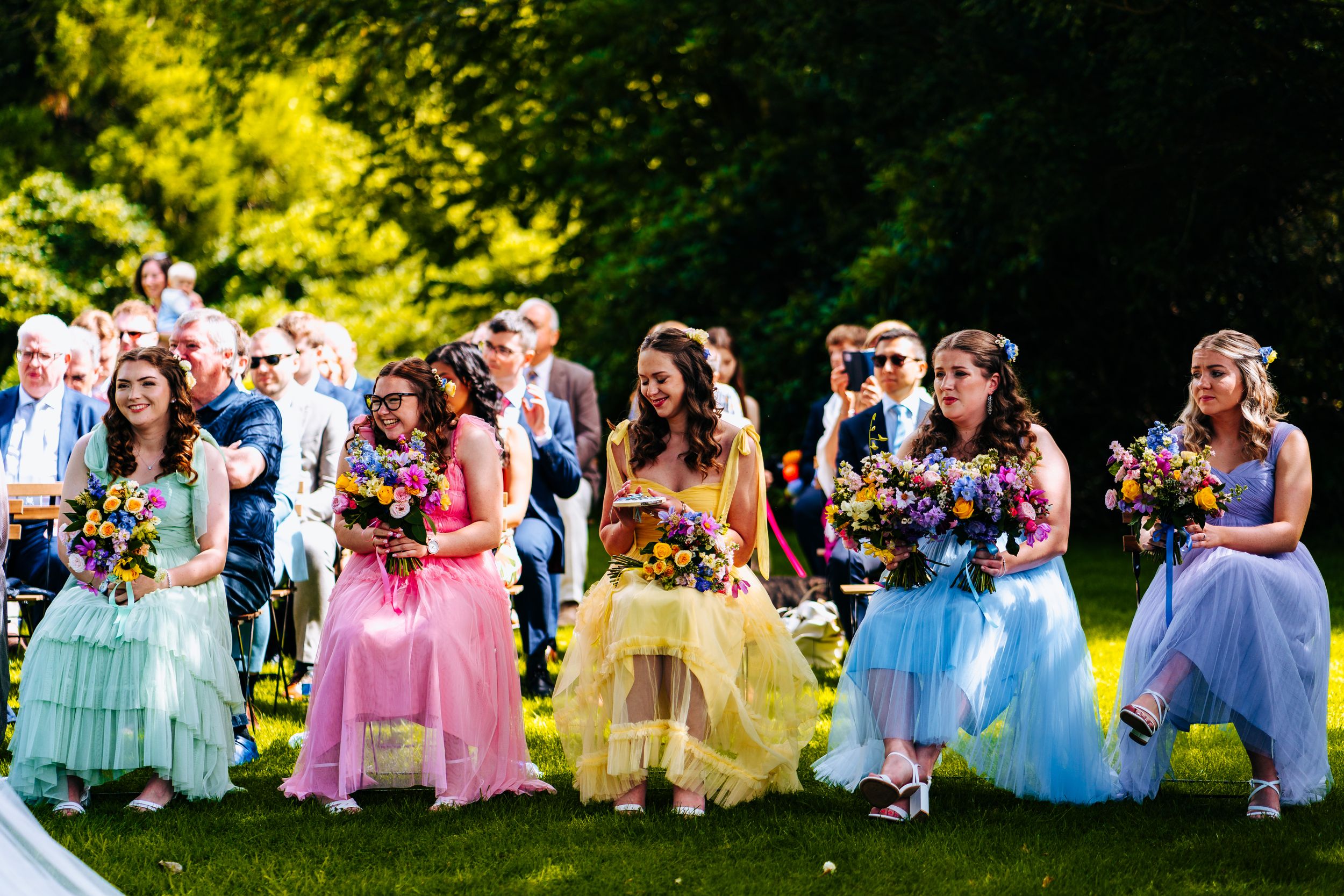 Bridesmaids walking in rainbow-colored tulle dresses carrying vibrant bouquets at outdoor wedding.