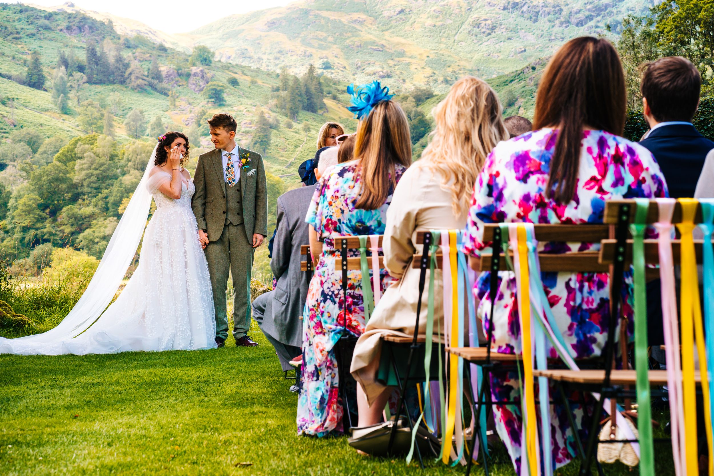 Bride and groom exchange vows at scenic outdoor wedding with lush green hills backdrop.