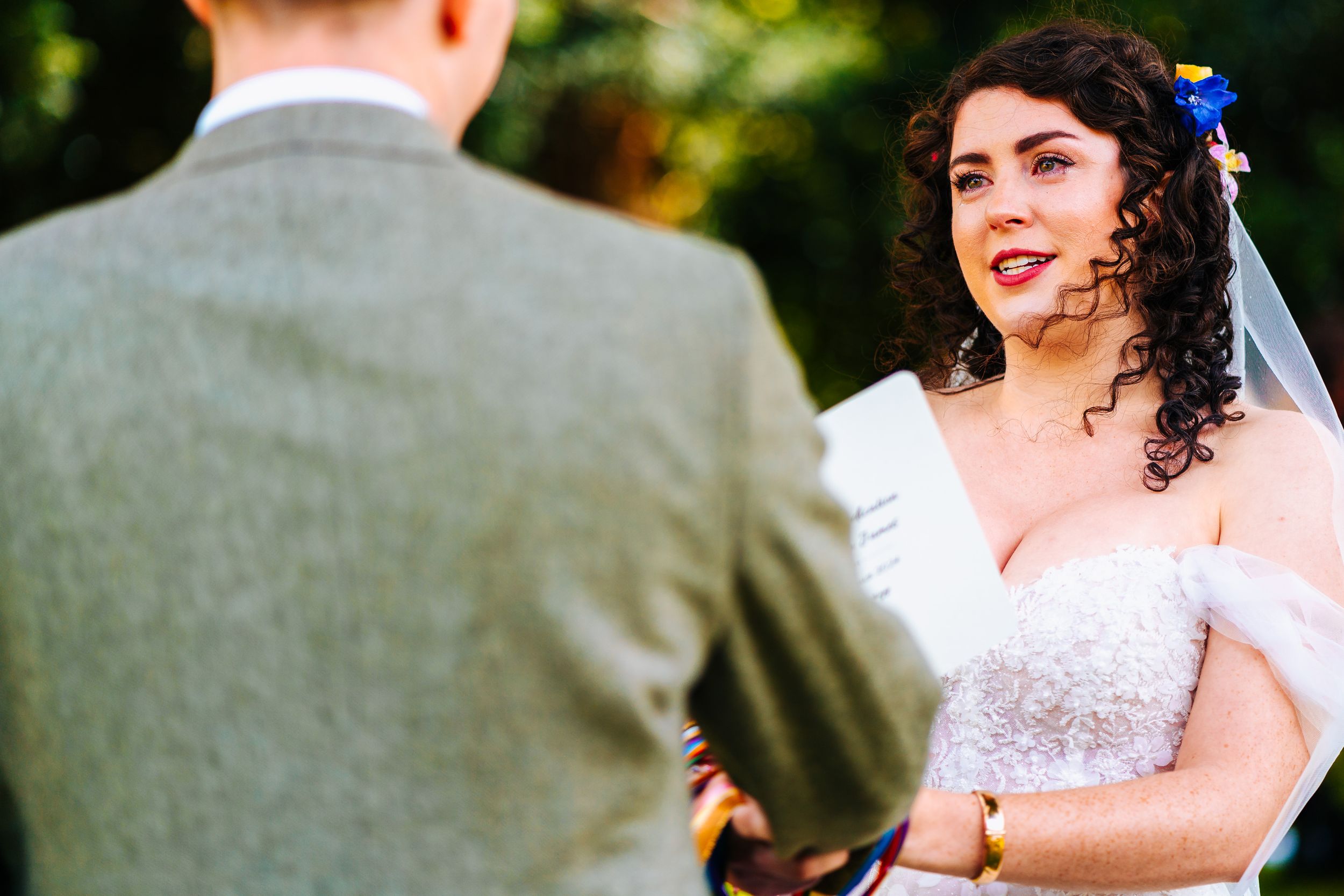 Bride in strapless lace wedding gown smiling joyfully at groom during outdoor wedding ceremony.