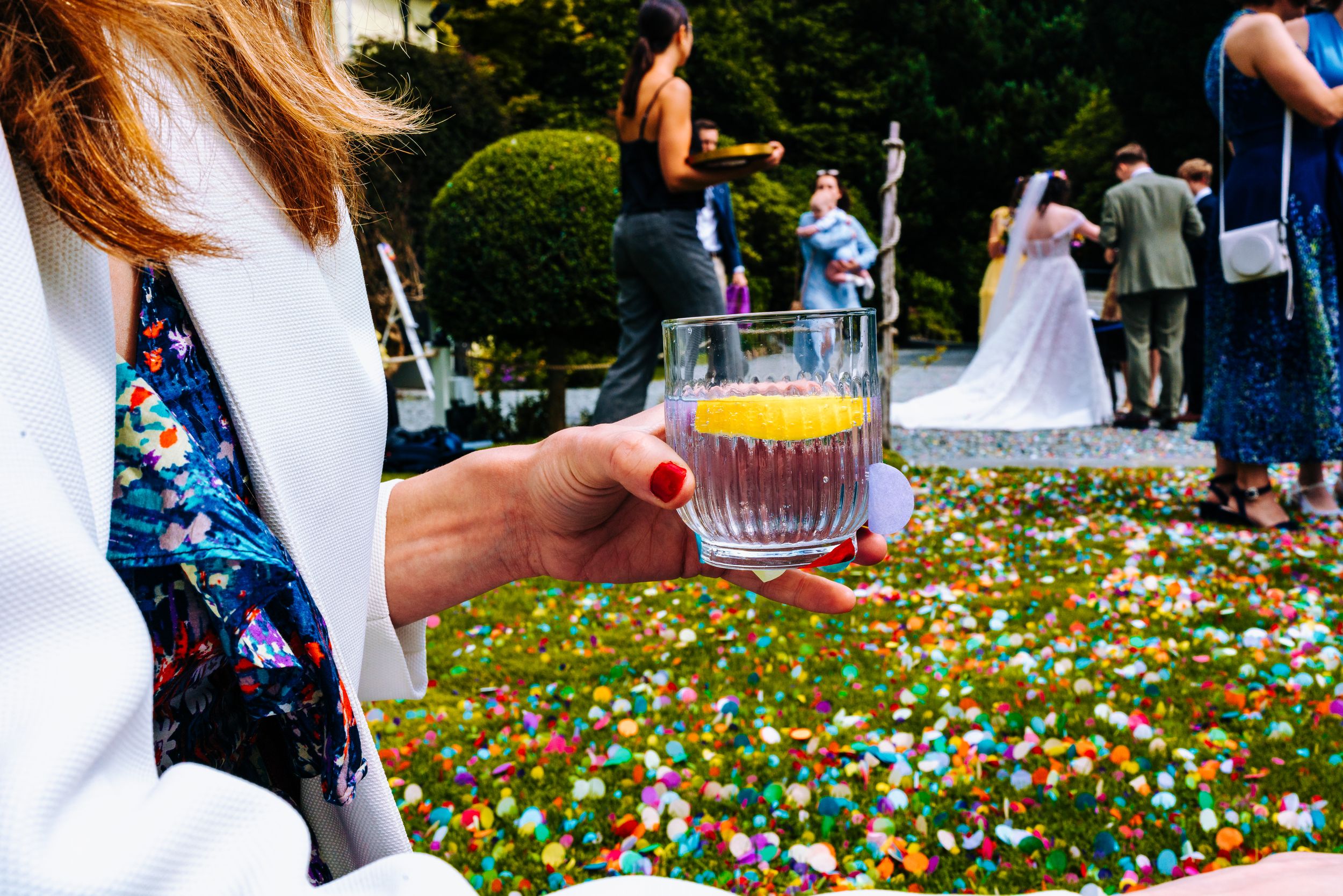 Guest holding crystal glass of orange juice at colorful confetti-covered wedding reception.