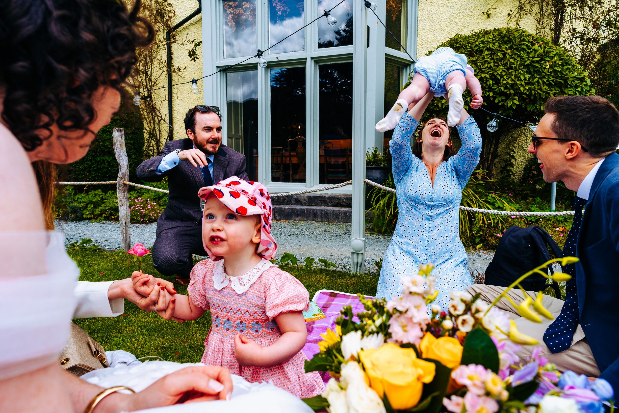 Wedding guests celebrating outdoors with couple lifting baby joyfully in garden setting.