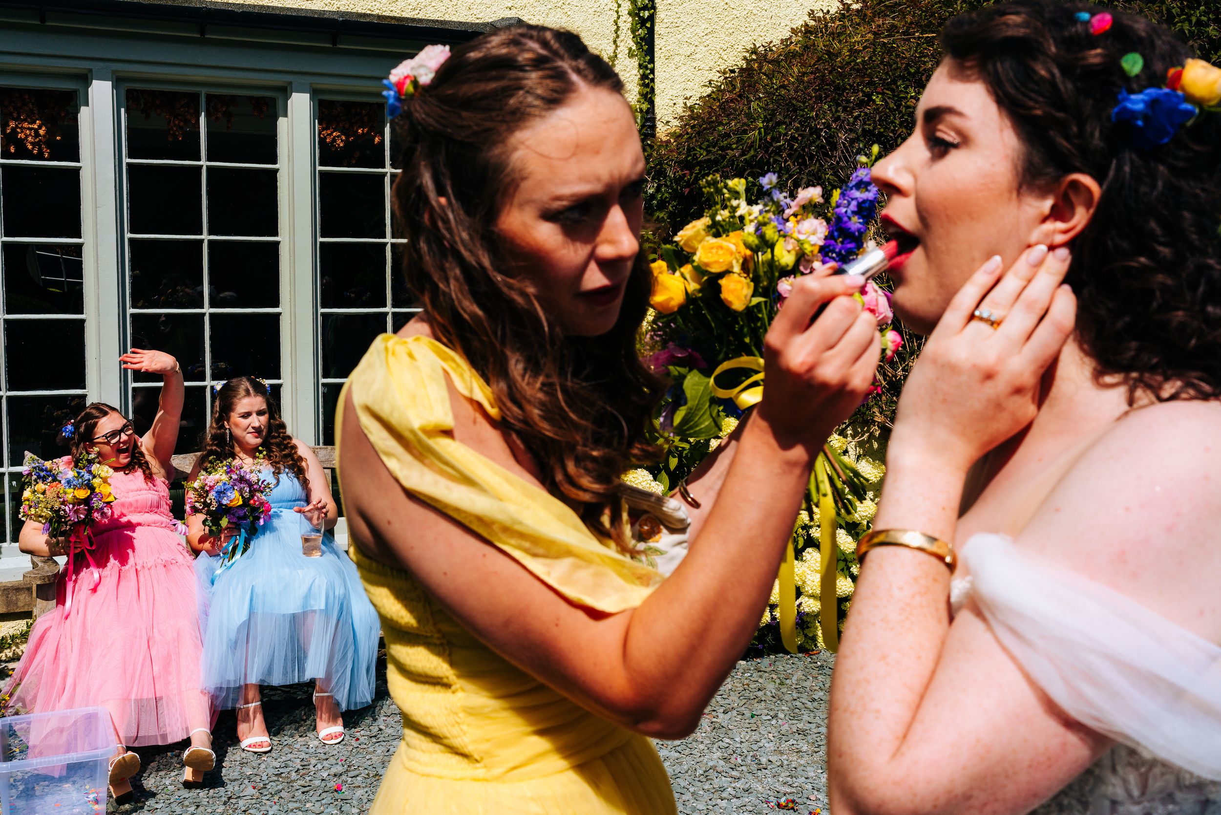 Bridesmaid in yellow dress applying lipstick to smiling bride with colorful hair accessories.