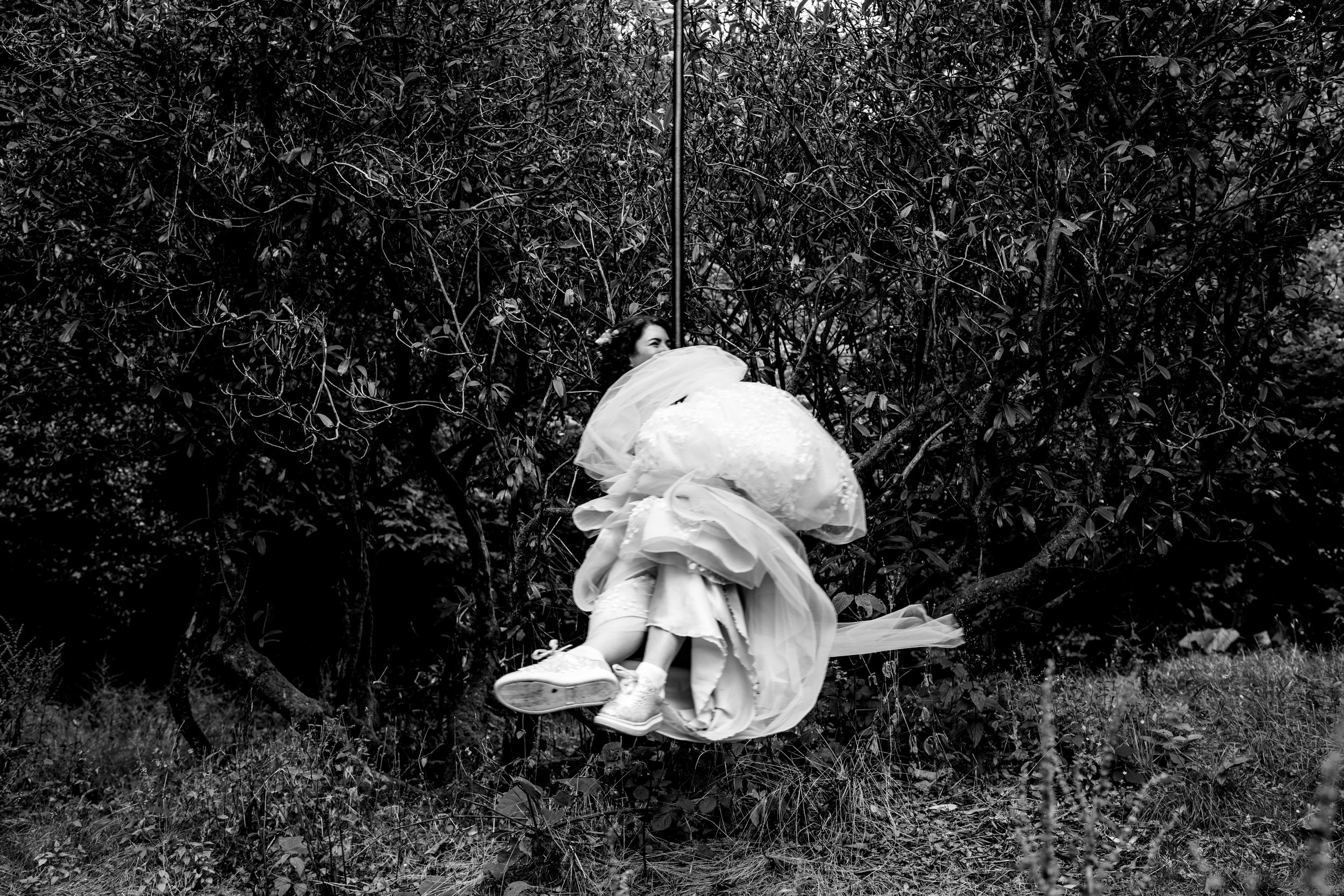 A child sits on a swing surrounded by lush green trees in a moody black and white outdoor photograph.