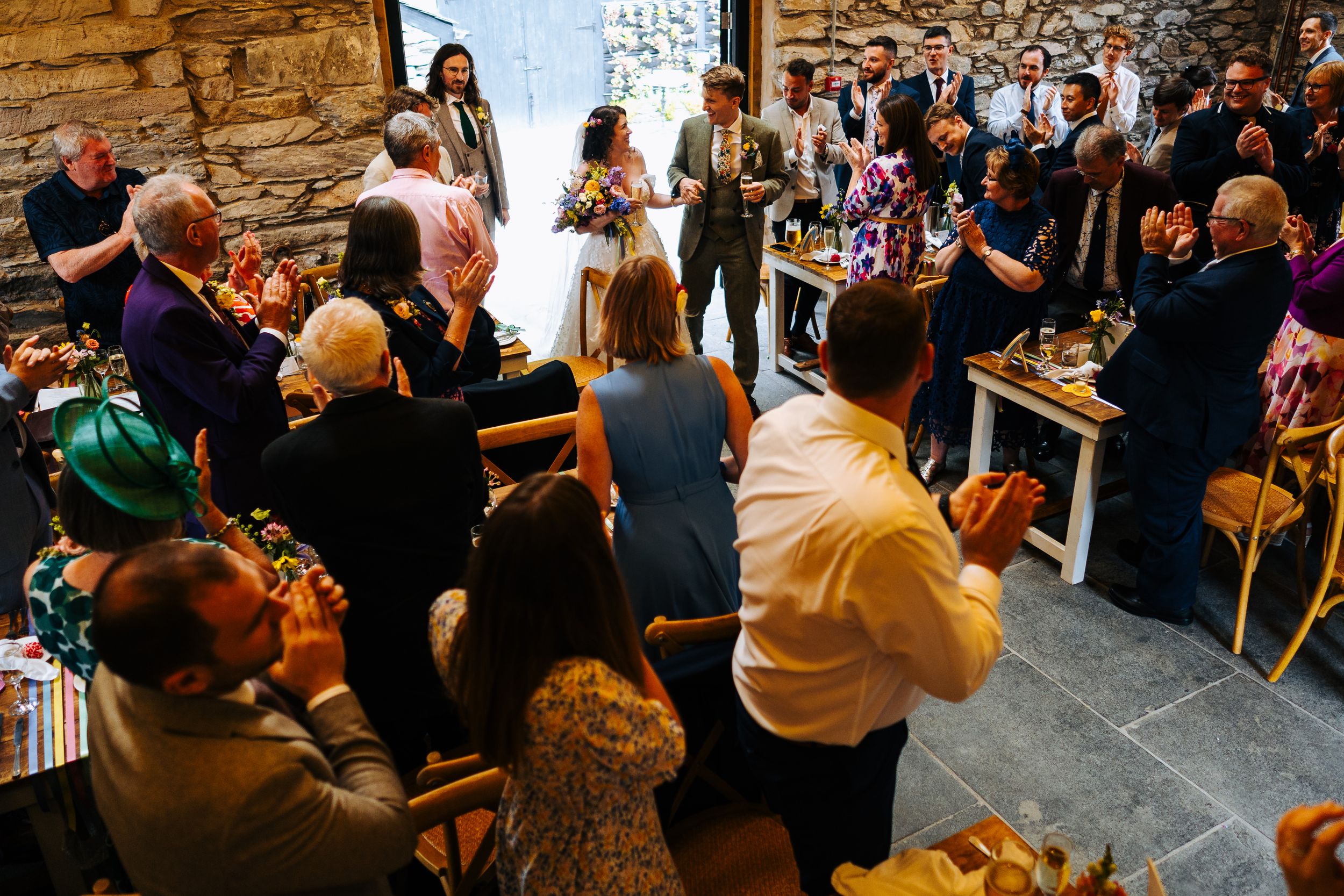 Wedding guests applaud the bride and groom during a reception in a rustic stone-walled venue.