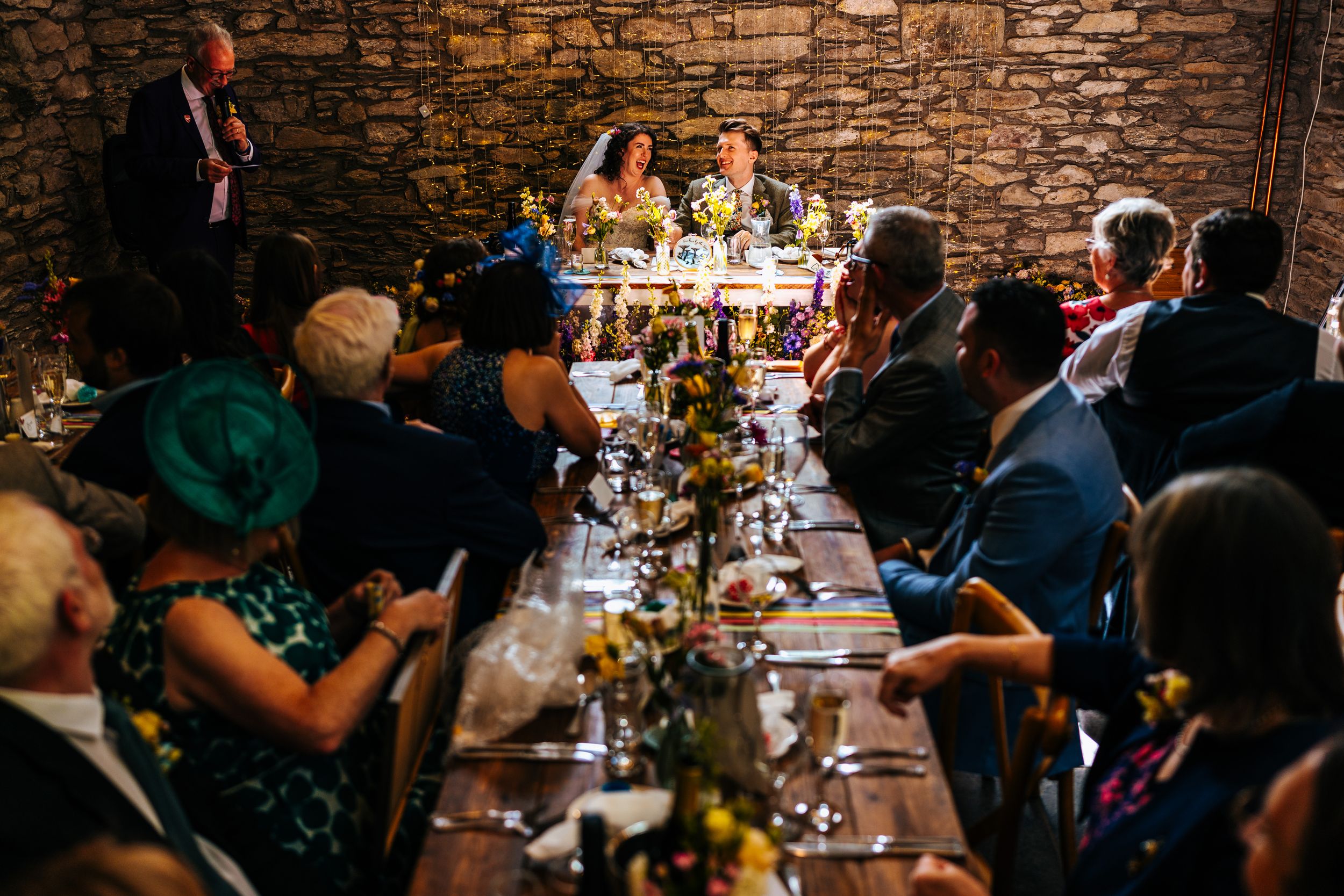 Wedding guests enjoy a reception dinner in a candlelit rustic stone barn with long decorated tables.