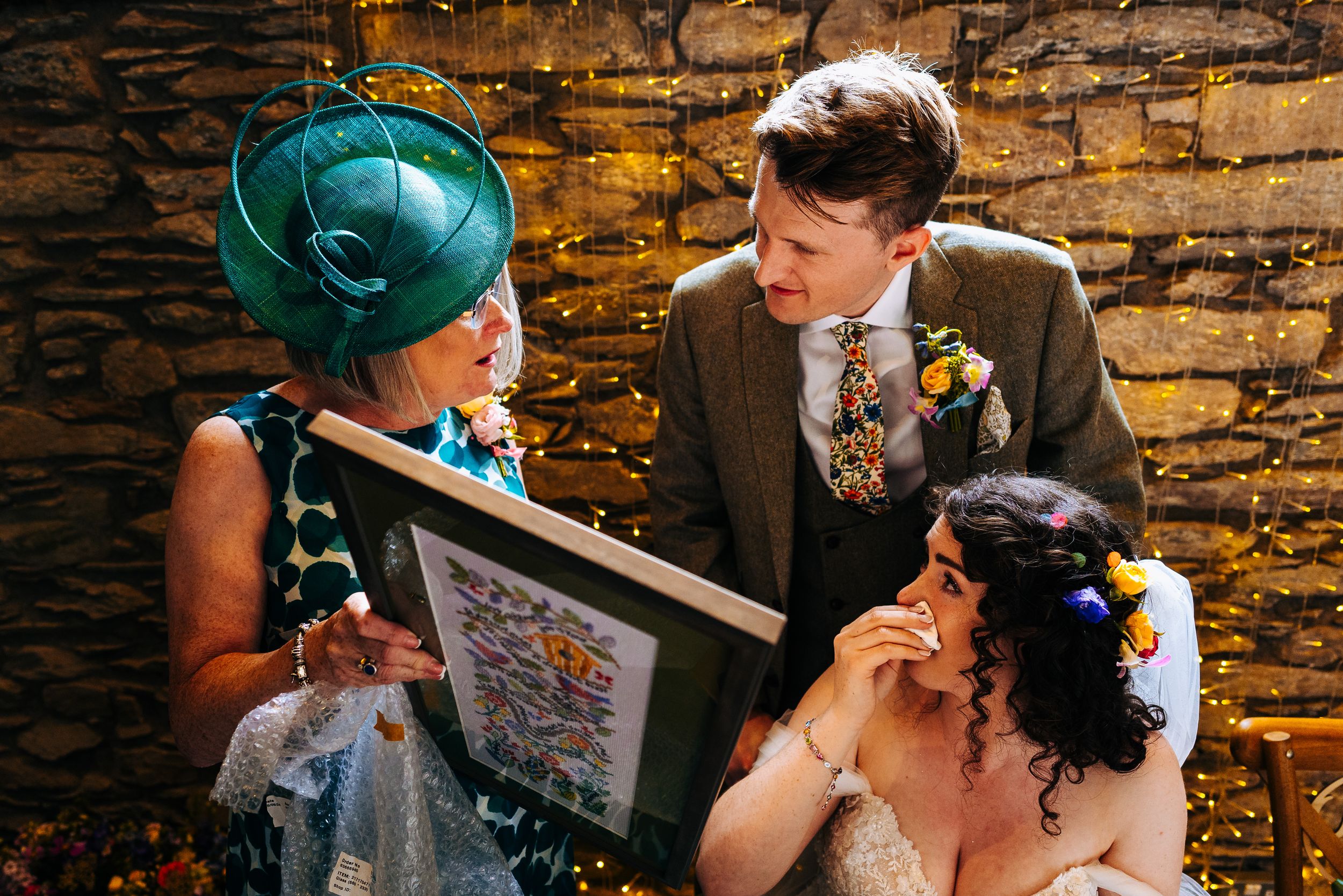 A bride and groom receive a framed gift from a guest wearing a teal fascinator at their wedding.