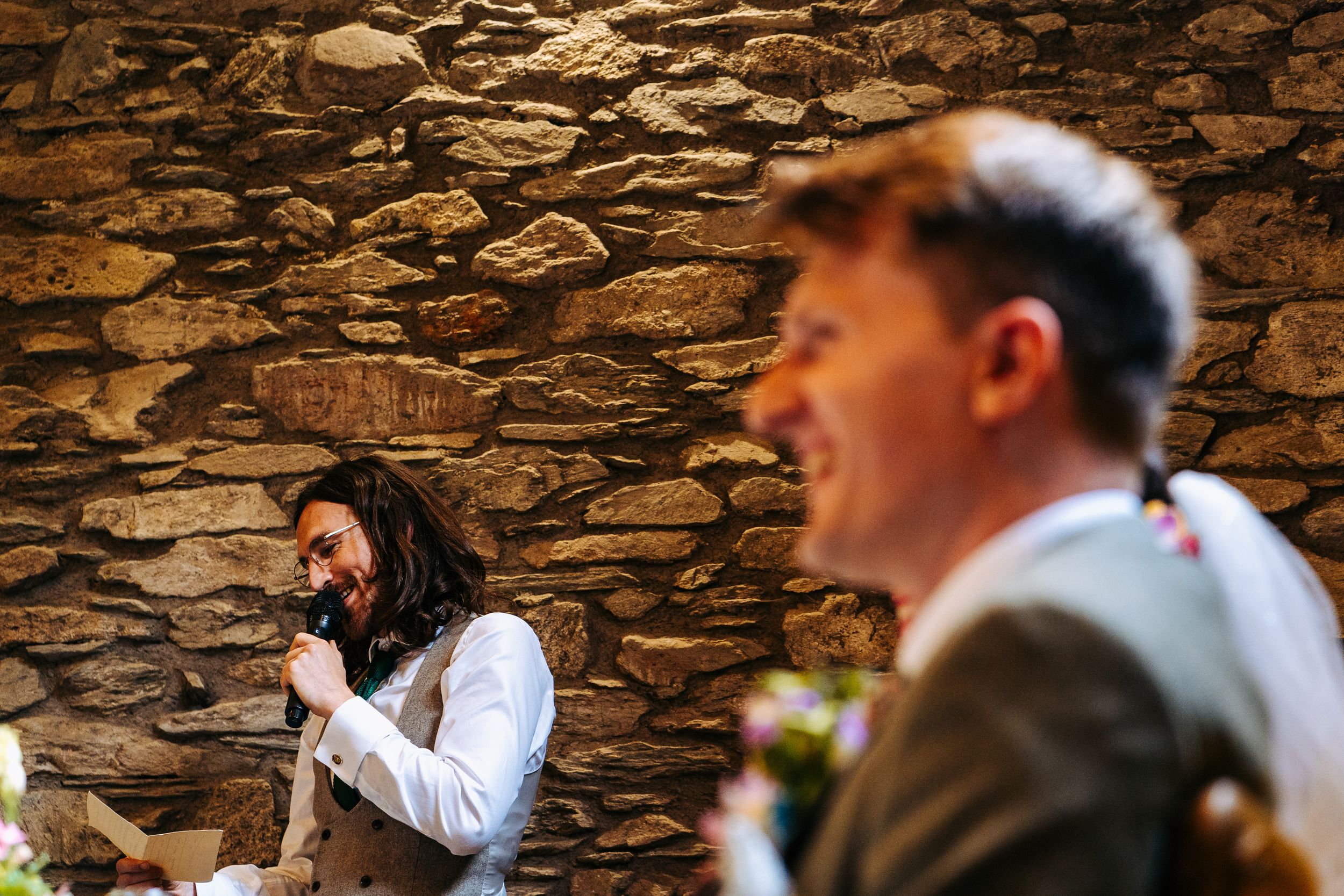 A wedding guest laughs candidly during speeches in a rustic stone-walled wedding reception venue.