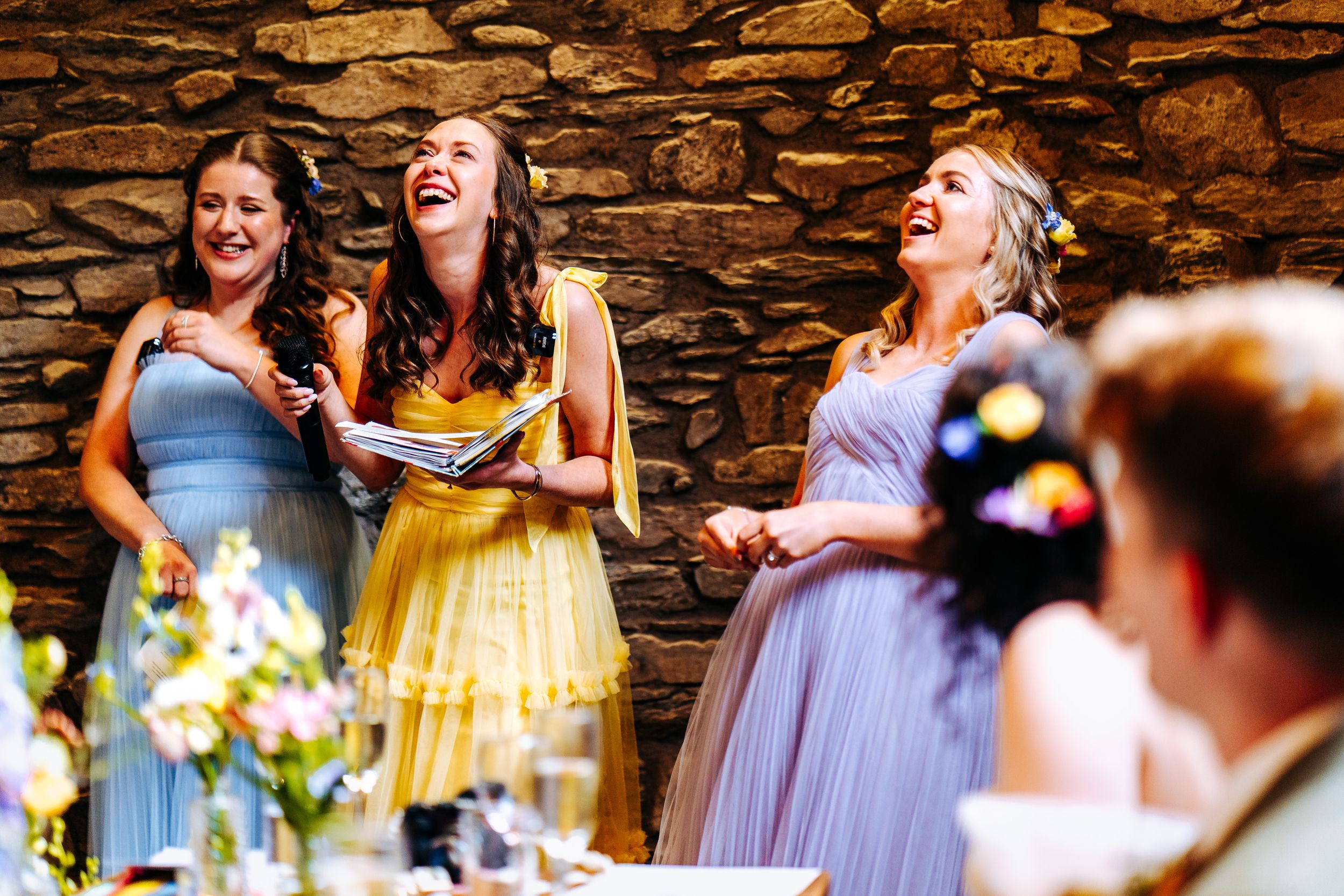 Three bridesmaids in colorful dresses laugh joyfully during wedding speeches at a rustic reception.