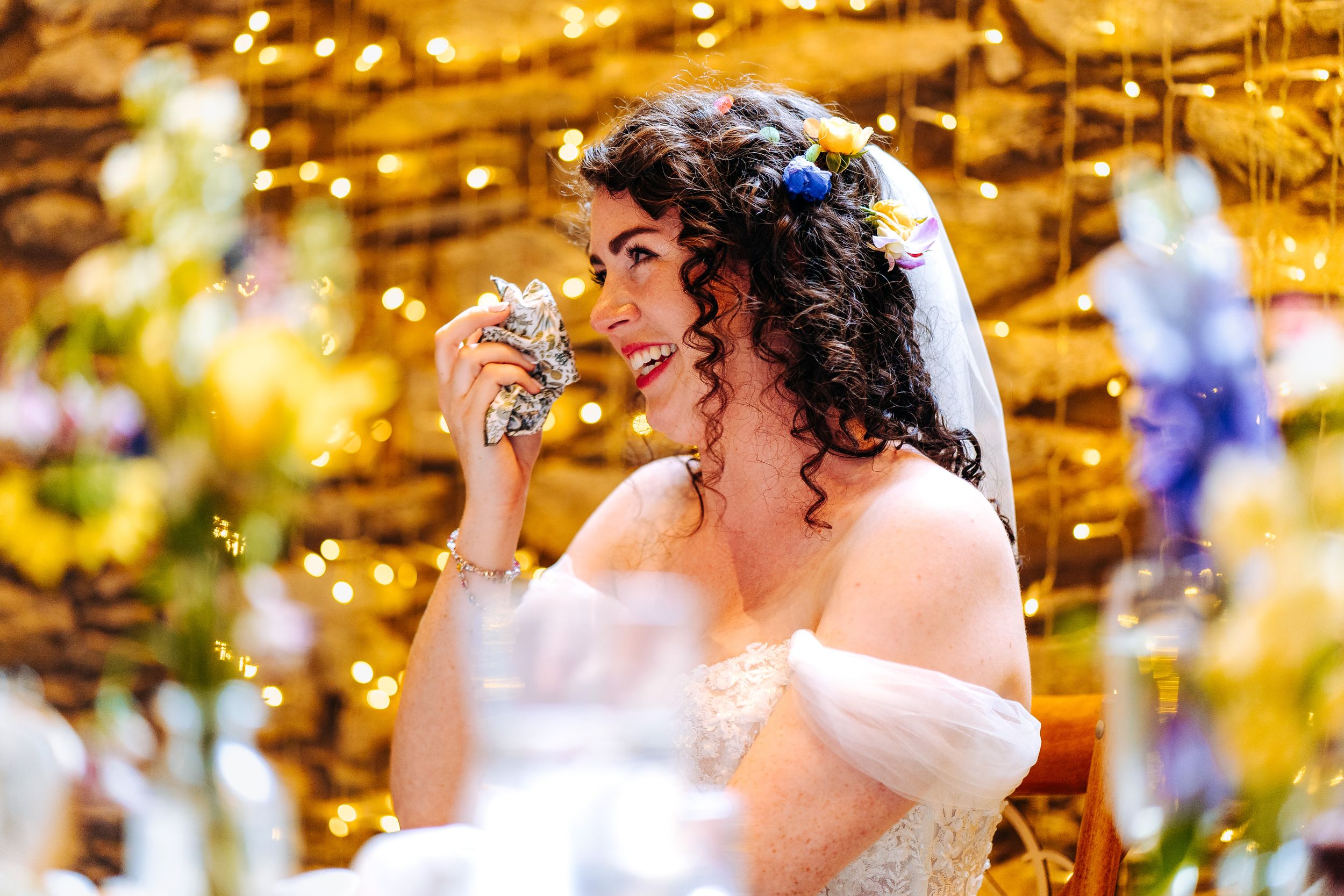 Radiant bride in white gown smiles holding a crystal glass amid warm fairy lights at her wedding reception.