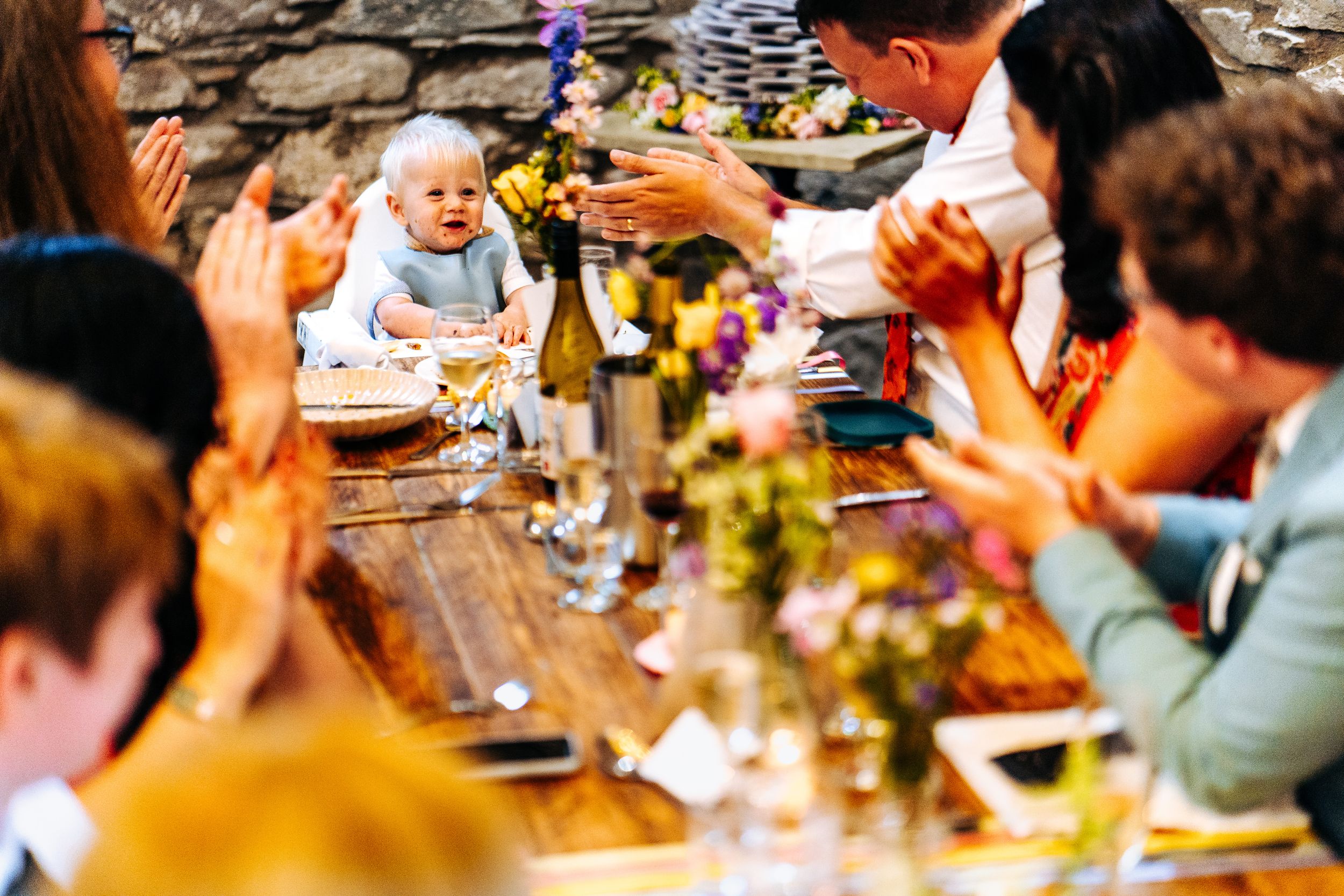 Baby sits at a rustic wedding reception table as guests clap and celebrate around floral centerpieces.