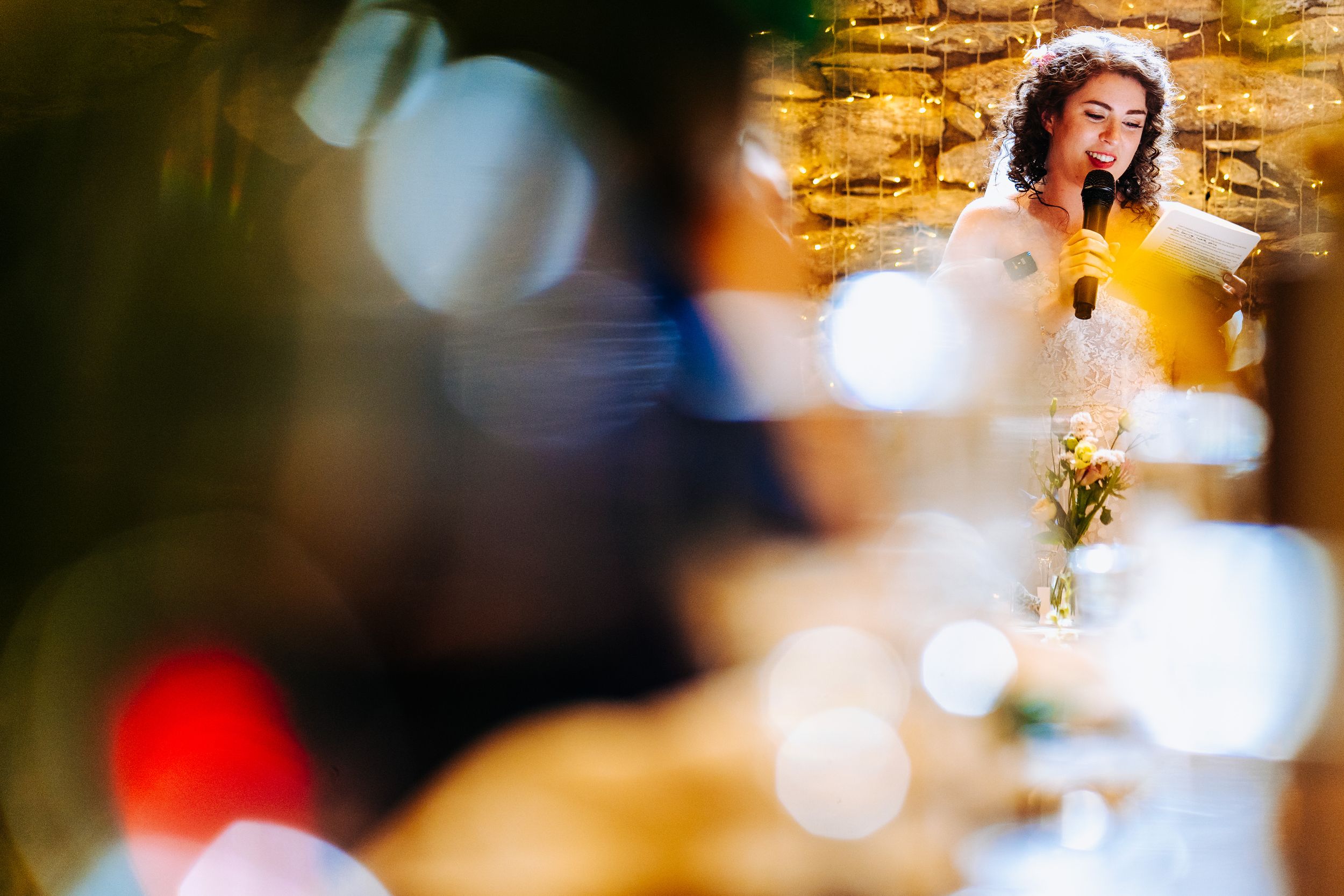Bride speaks into a microphone at the wedding reception captured through a dreamy bokeh foreground.