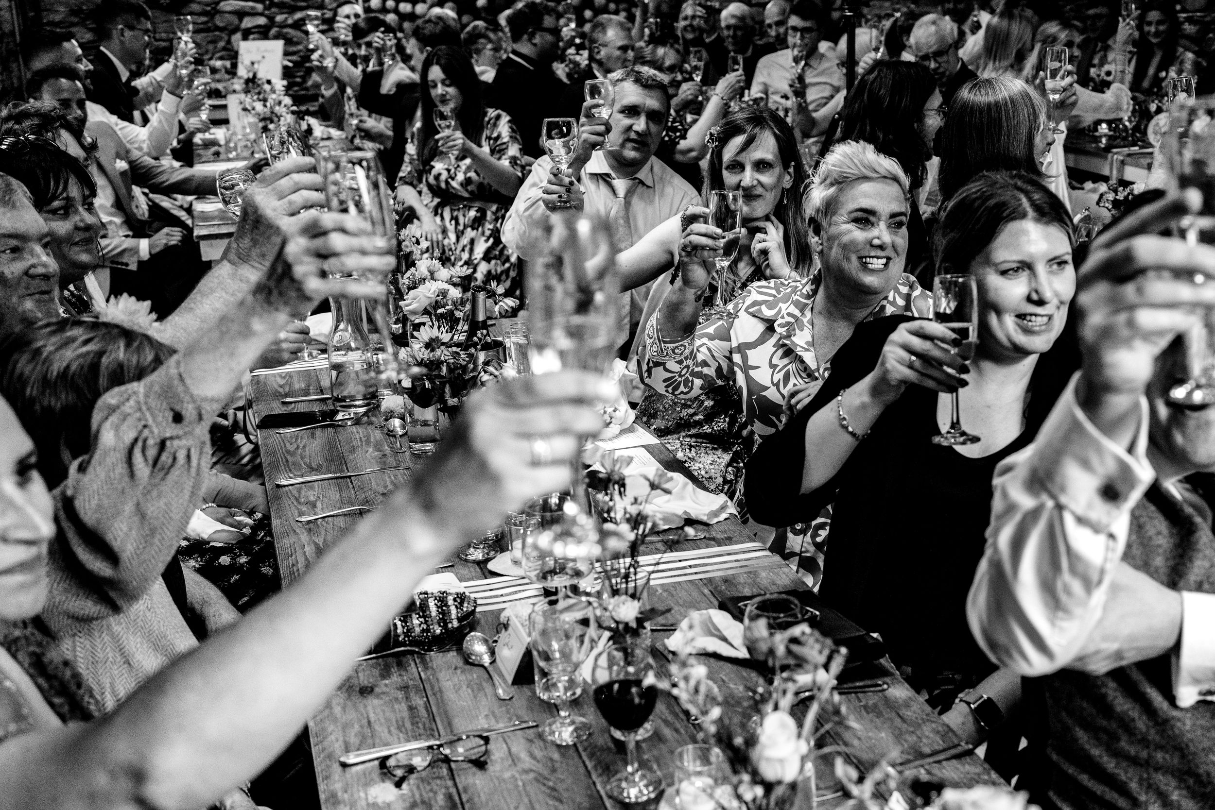 Wedding guests raise glasses in a joyful toast at a long rustic table in black and white photo.