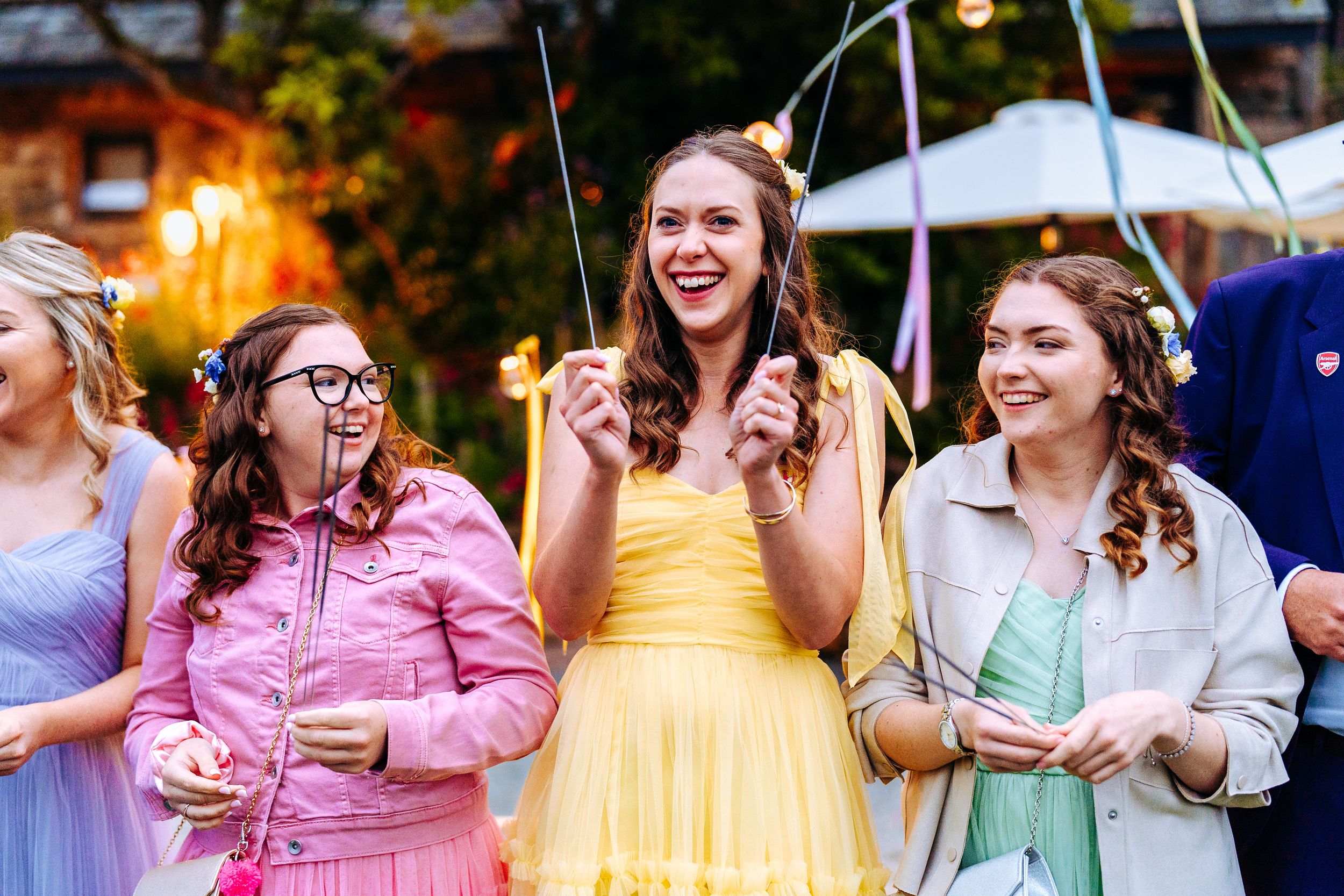 Three smiling women in colorful dresses hold sparklers at an outdoor evening wedding celebration.
