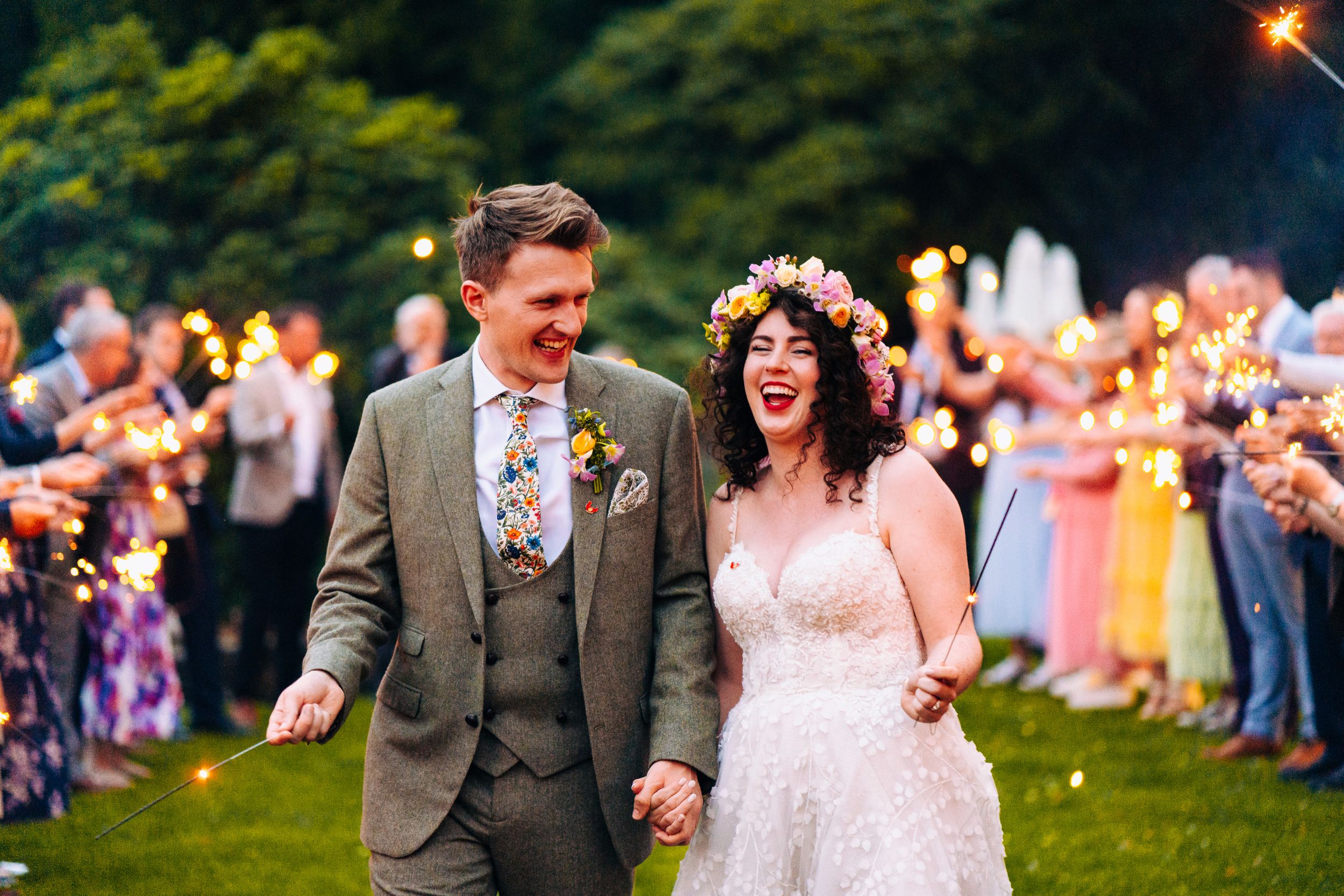Bride wearing floral crown and groom walk hand in hand through a sparkler tunnel at their wedding.