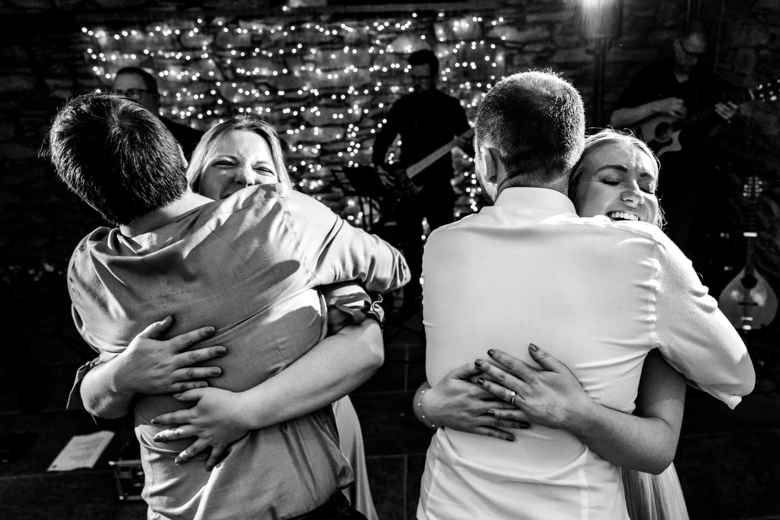 Two couples share warm embraces on the dance floor in an emotional black and white wedding photo.