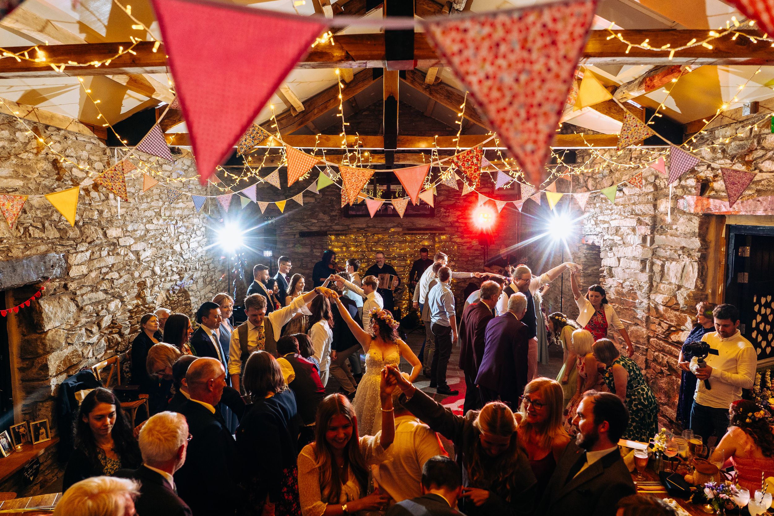 Guests dancing and celebrating at a rustic wedding reception with colorful bunting and fairy lights.