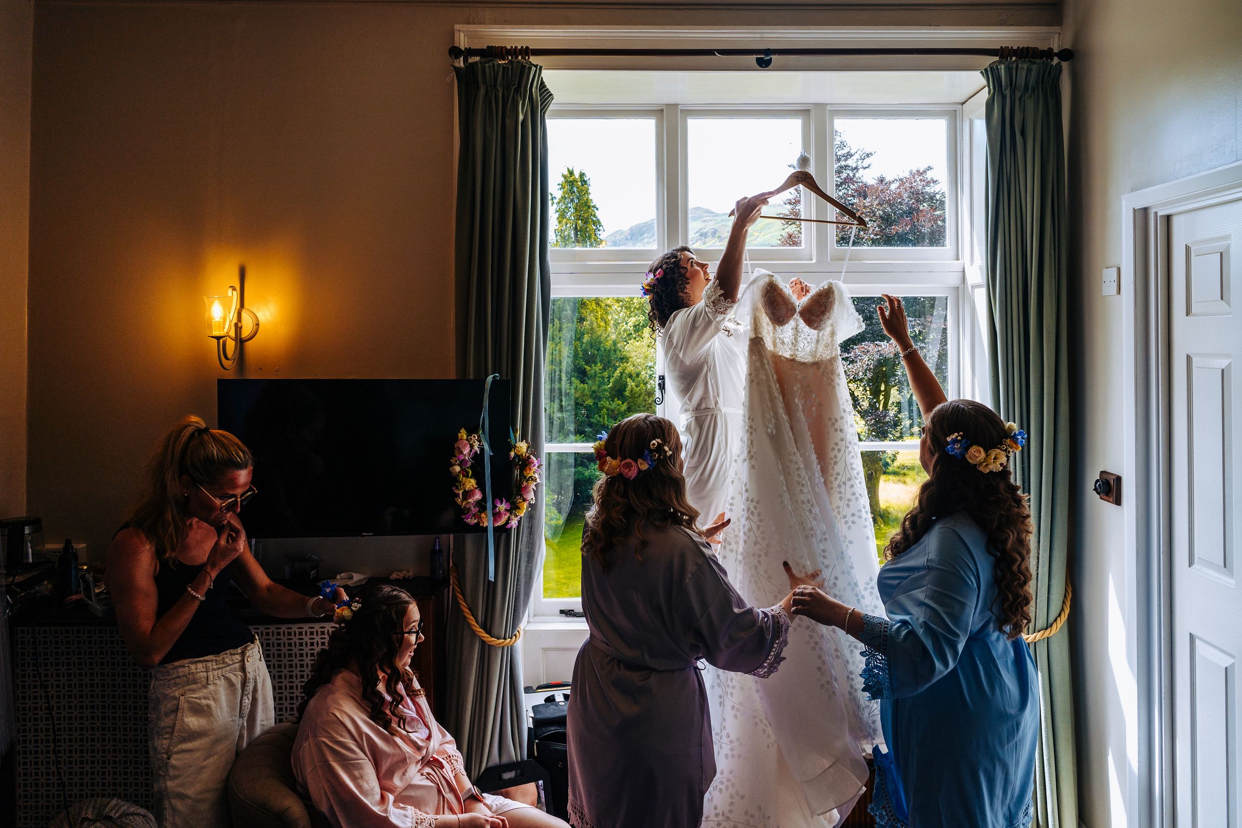 Bridesmaids helping lift a wedding dress by a window in a warmly lit bridal suite at the Lancrigg in Grasmere