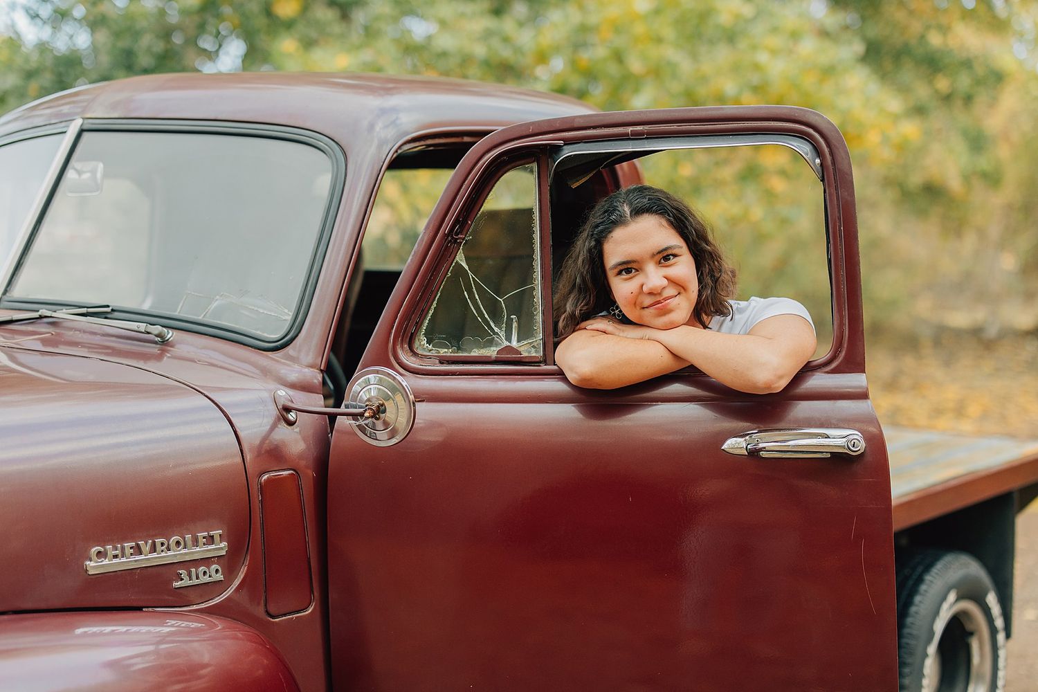 Smiling young woman leans out window of vintage red Chevrolet truck in autumn setting.