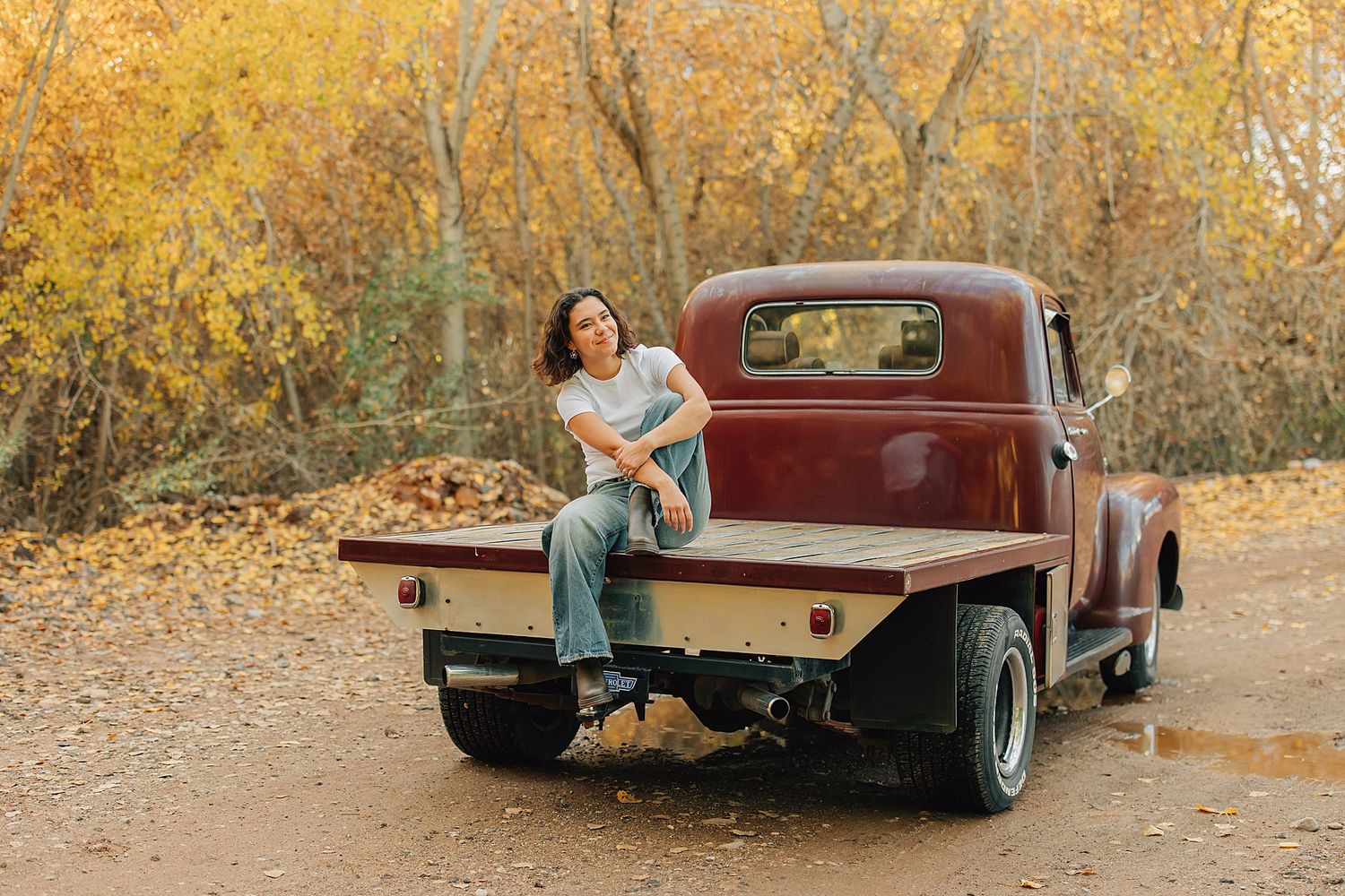 Woman in jeans sitting on the flatbed of a vintage red pickup truck surrounded by autumn foliage.