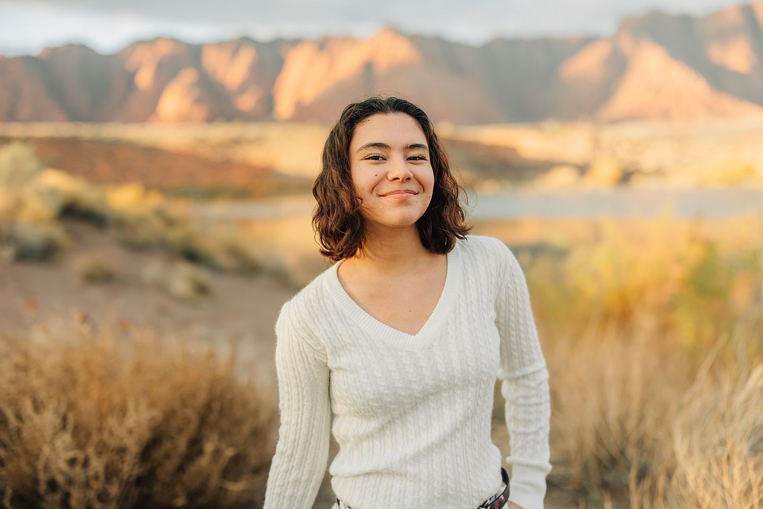 Young woman in white sweater smiling outdoors with desert mountains and golden grasses in warm sunset light.