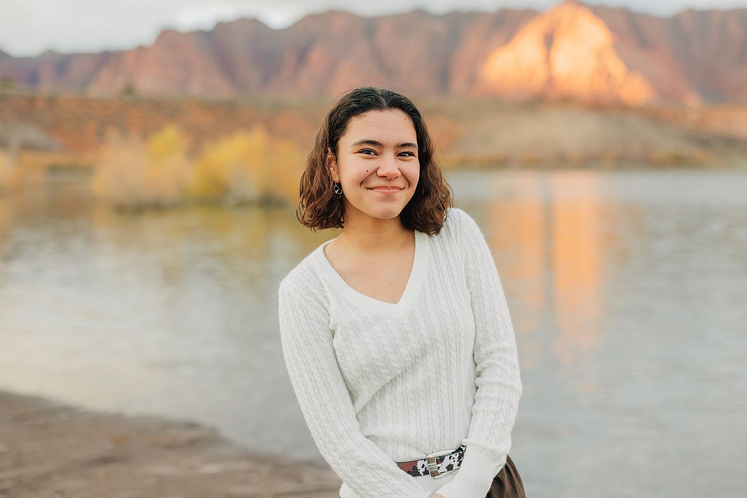 Young woman in white sweater smiling outdoors near a river with red rock formations in the background.