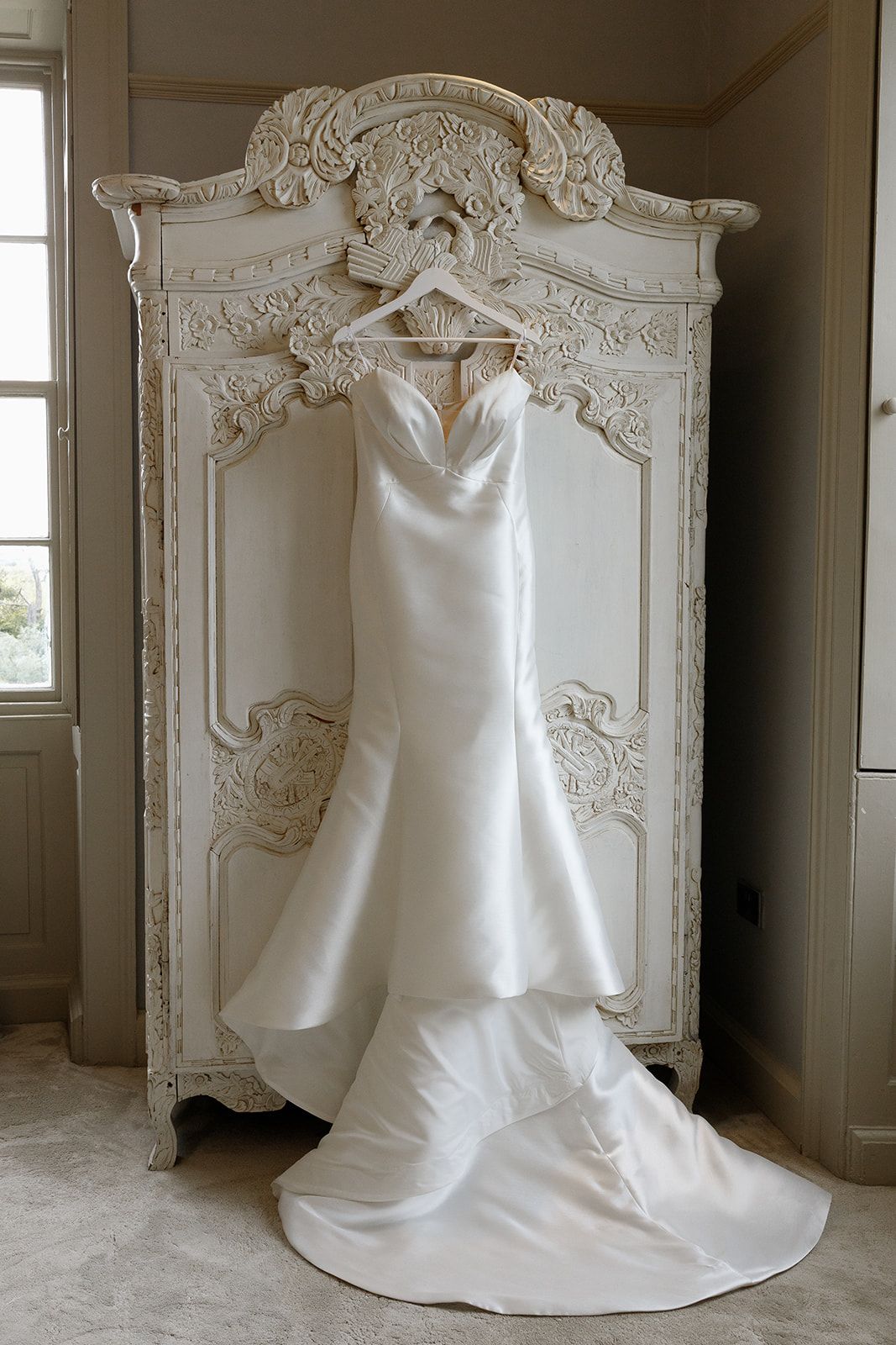 Elegant white satin mermaid wedding dress hanging on ornate vintage armoire in soft natural light.