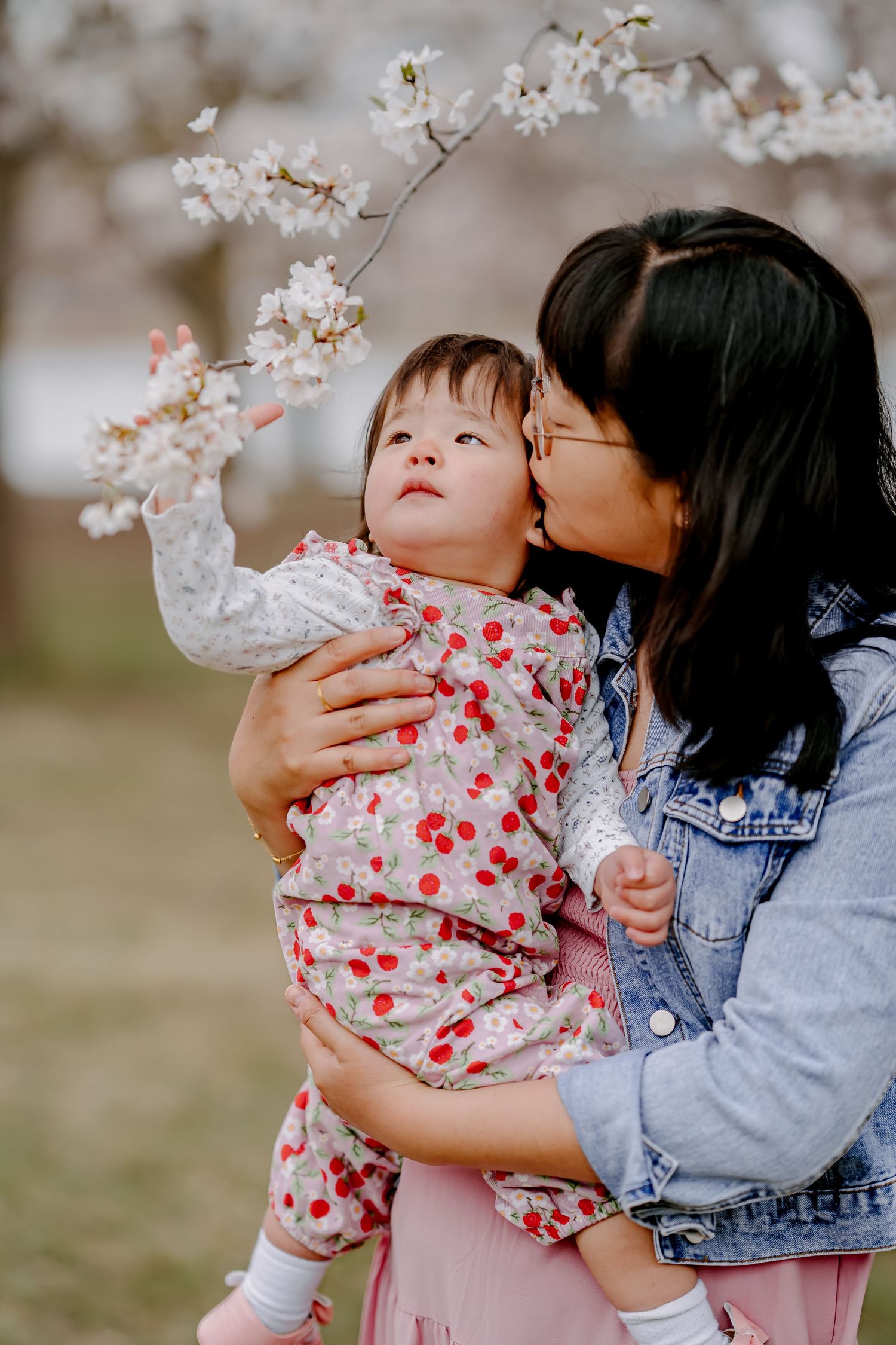 A mother kisses her toddler girl in a cherry blossom outfit while standing under white flowering trees in spring.