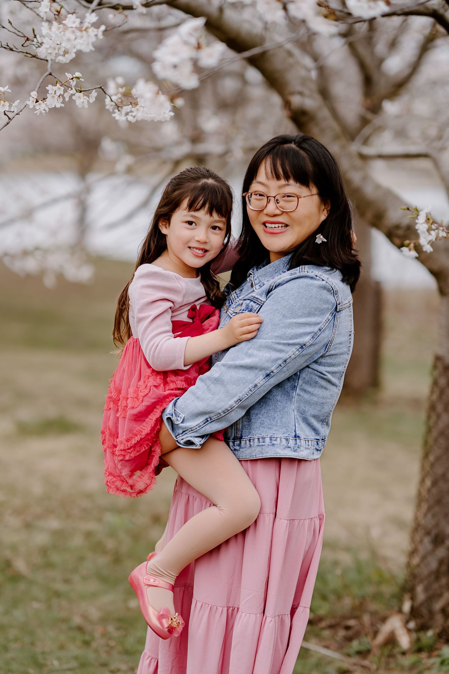 A mother holds her young daughter among blooming cherry blossom trees, both wearing pink outfits in a spring garden.