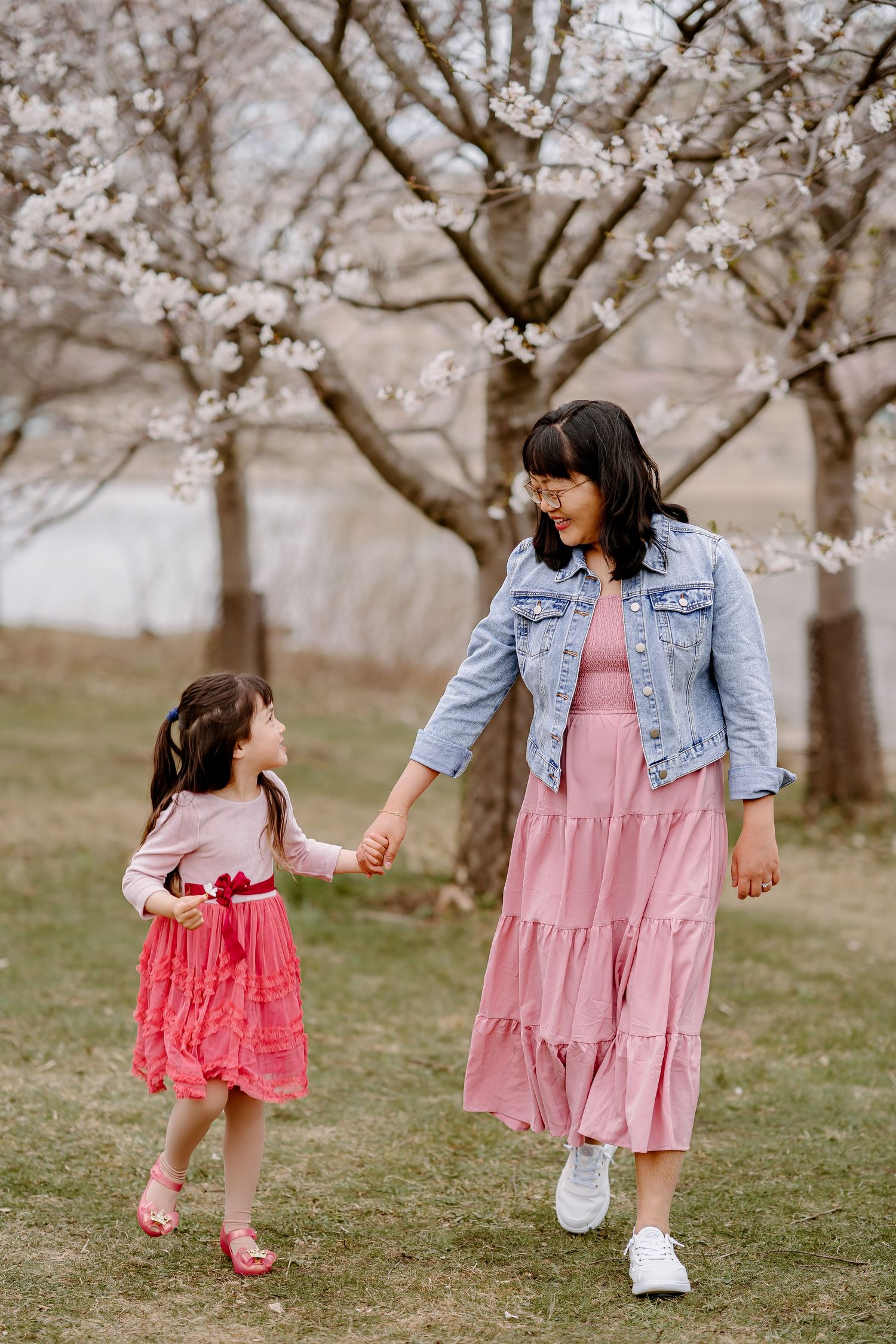 A mother and daughter in matching pink outfits hold hands while walking among blooming cherry blossom trees.