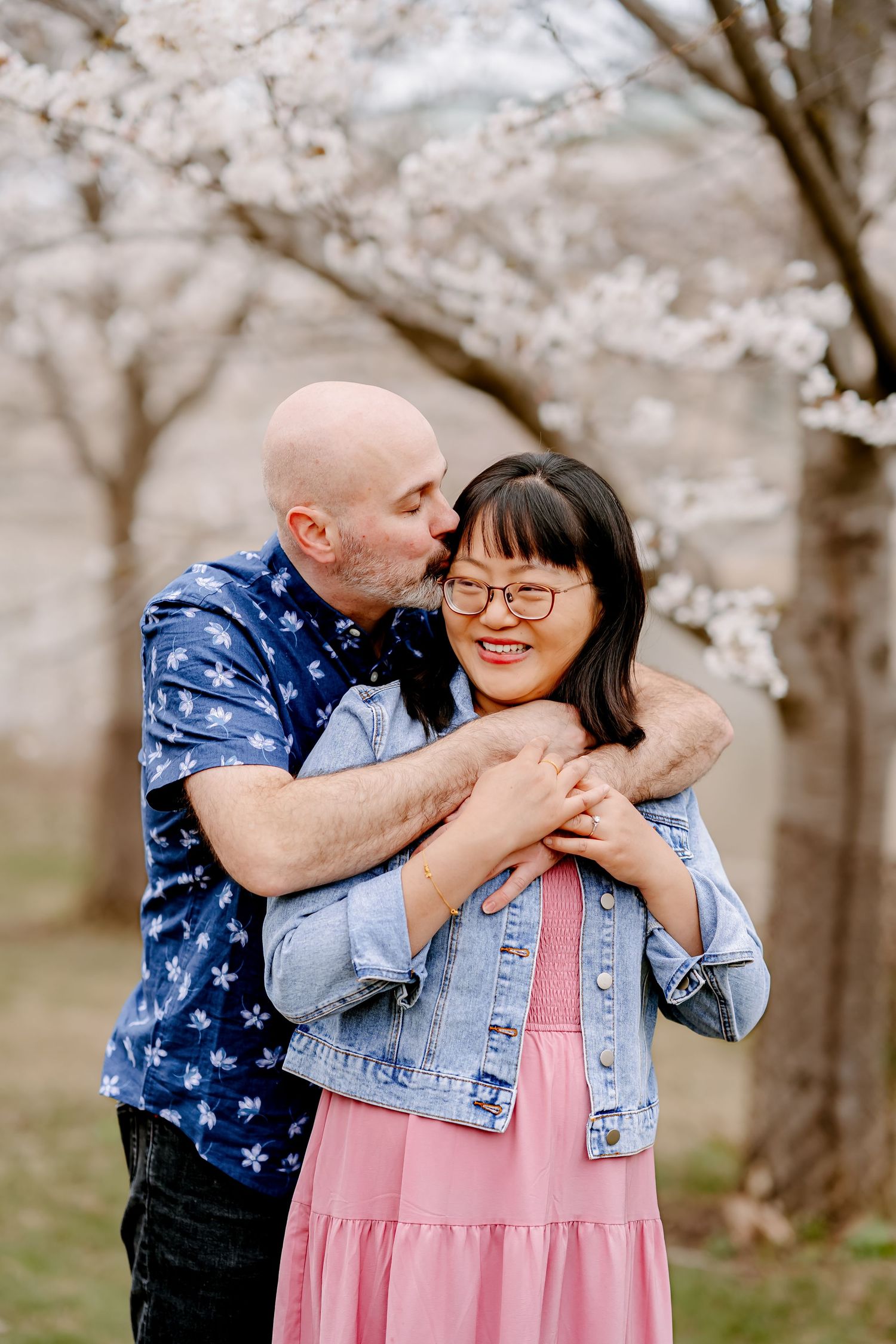 A bald man embraces a smiling Asian woman wearing glasses near cherry blossom trees in spring.