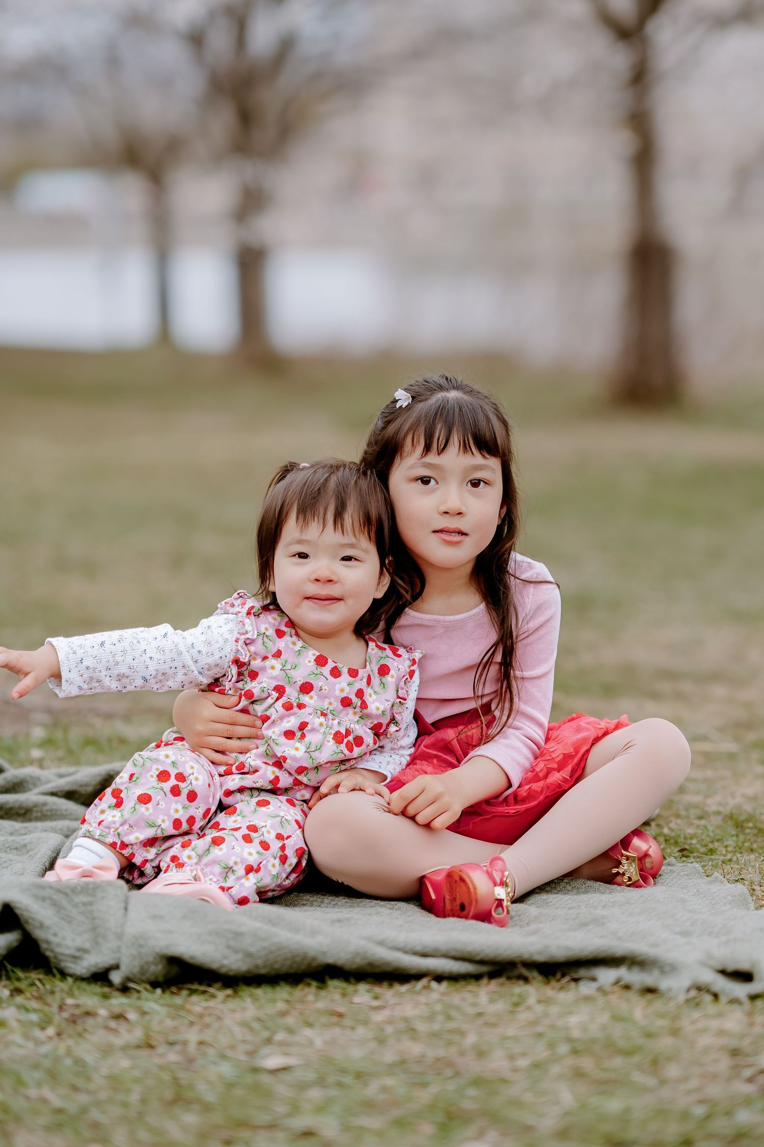 Two young Asian sisters sitting on a blanket in a park, older girl hugging younger toddler, both smiling at camera.