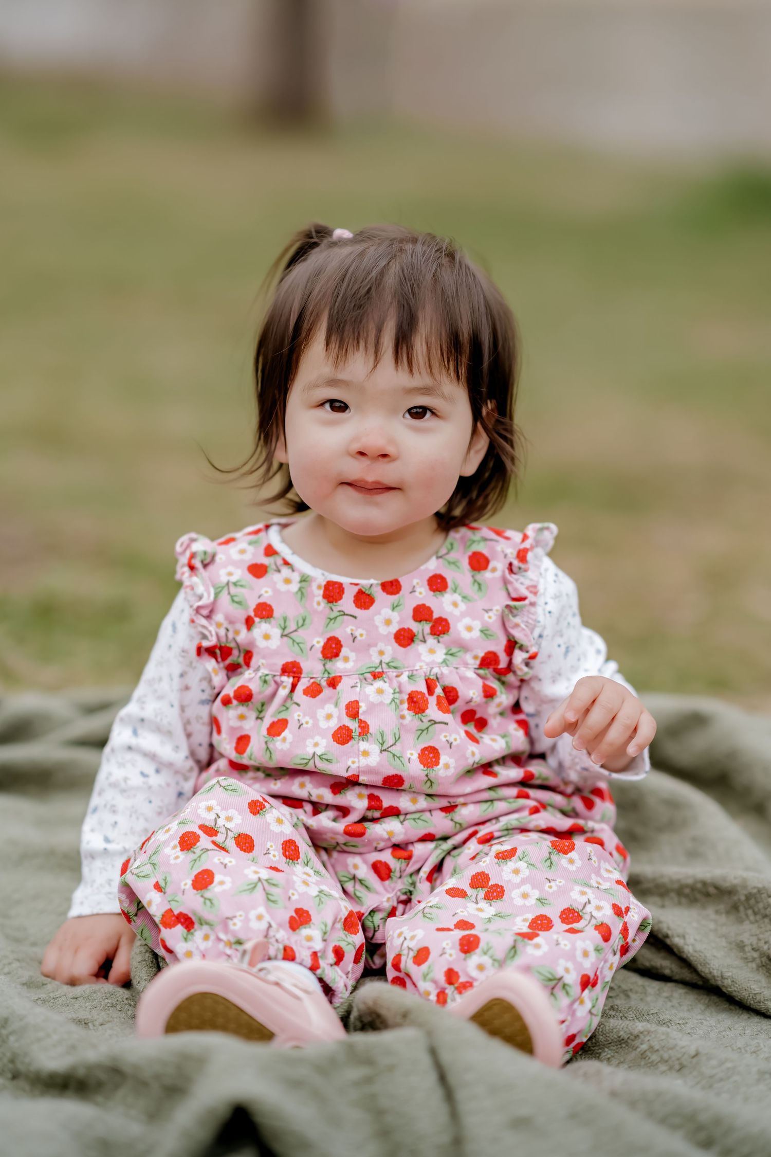 Adorable baby girl in cherry print outfit sitting on green blanket outdoors, smiling at camera.
