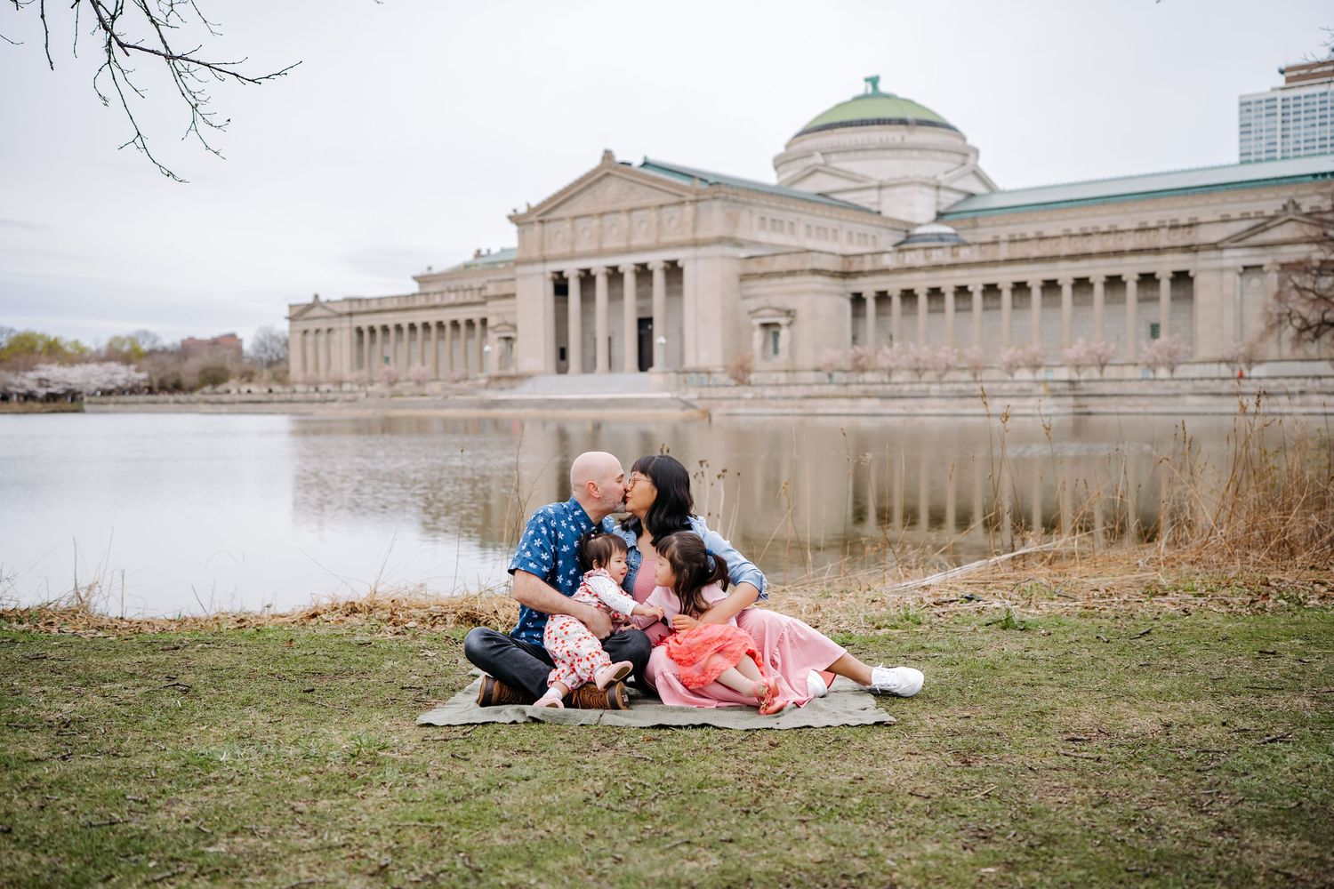 Family sitting together on a blanket by a pond near a neoclassical museum building on an overcast day.