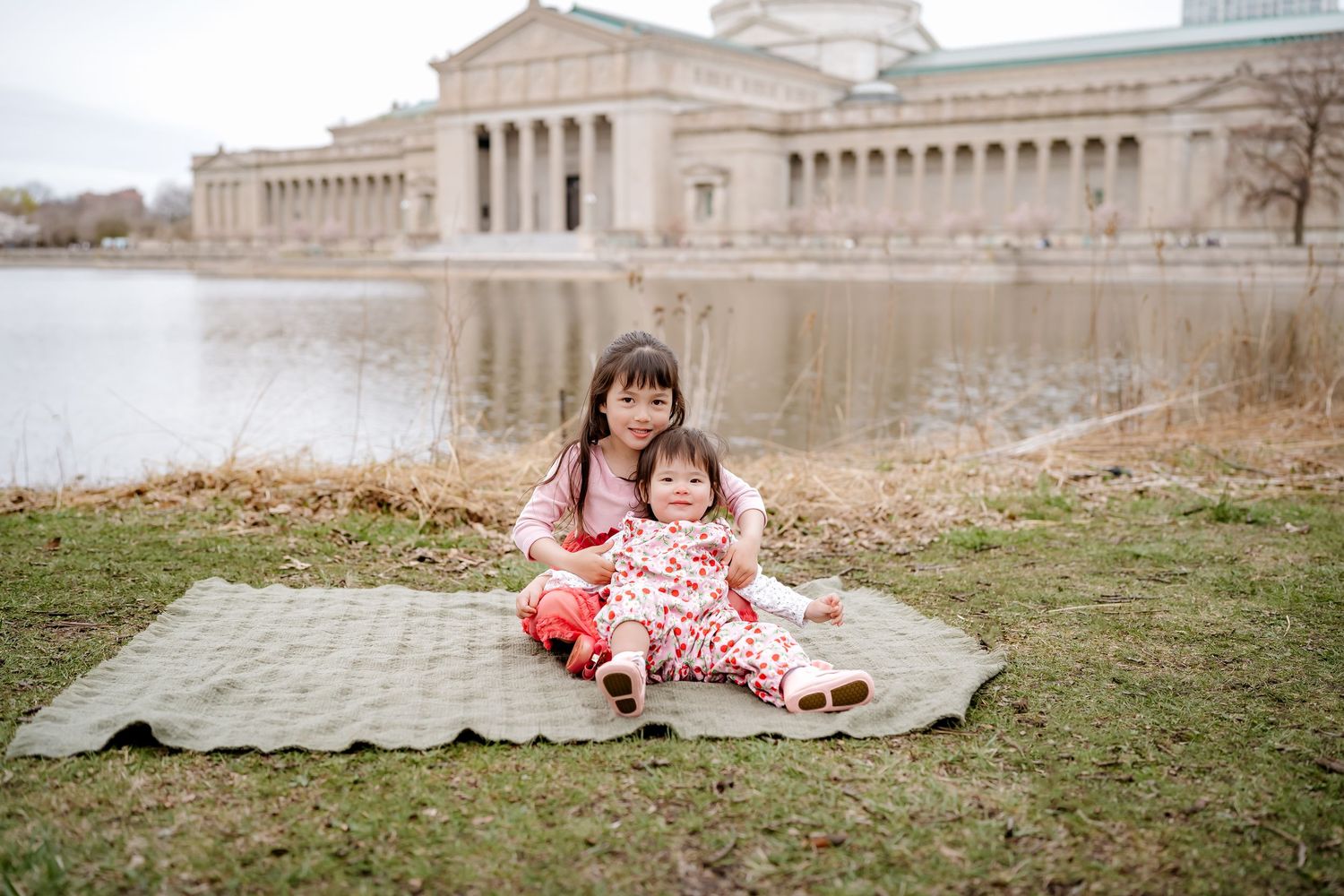 Two young sisters sitting on a blanket by a pond near a classical museum building outdoors.